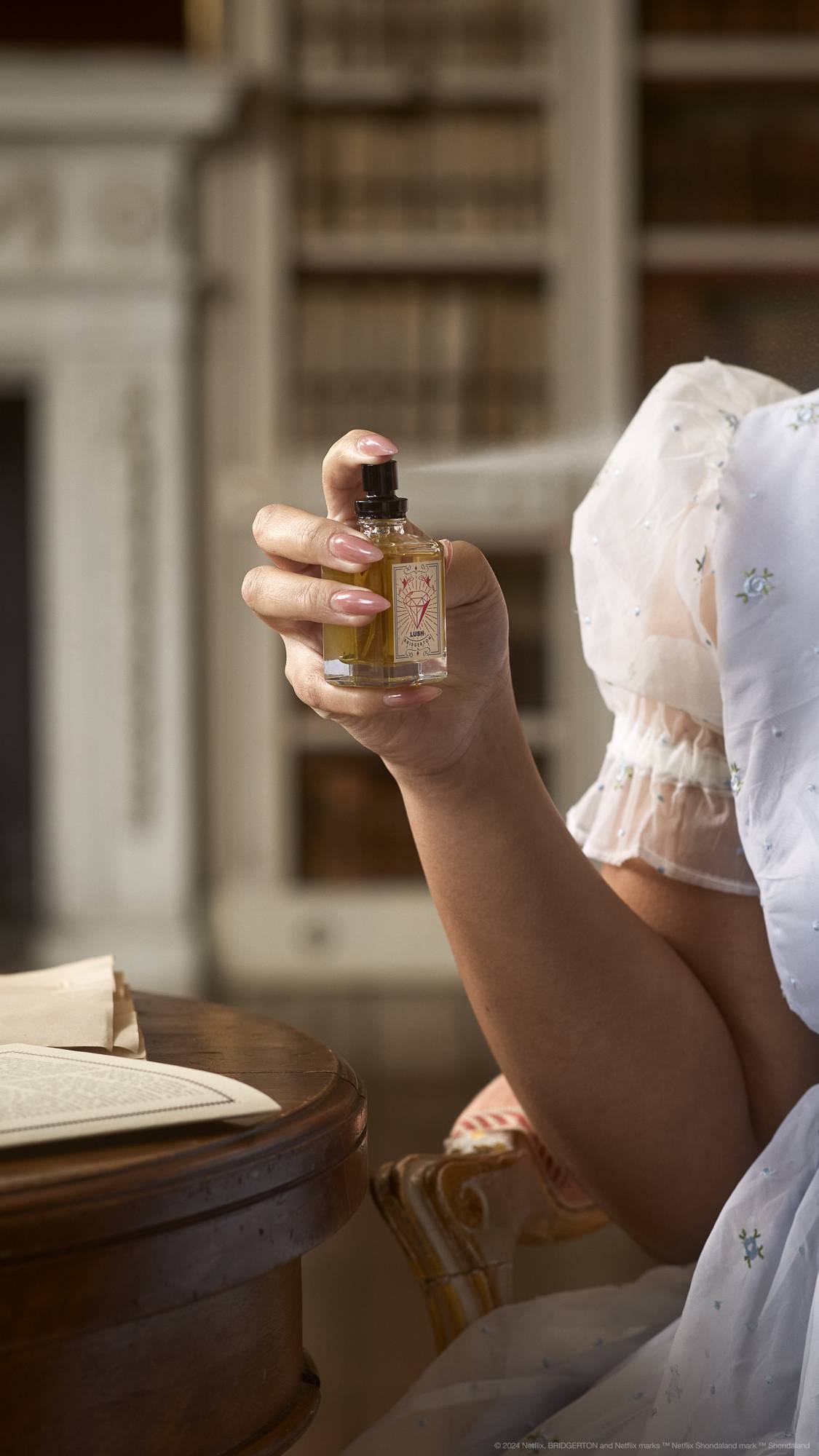 A close-up image of the model's hand holding the LUSH | Bridgerton perfume bottle as they are-mid spritz.