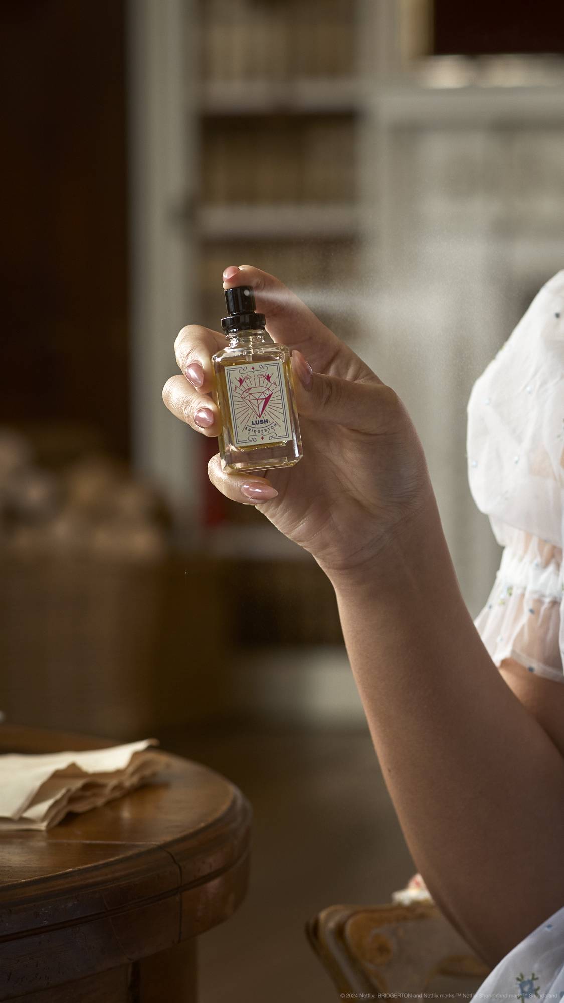 A close-up image of the model's hand holding the LUSH | Bridgerton perfume bottle as they are-mid spritz.
