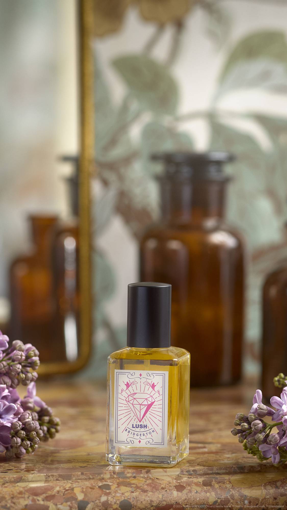 An out-of-focus background of leaves and what appears to be a dressing table. The focus is the LUSH | Bridgerton perfume bottle placed on a marbled counter with flowers on either side.
