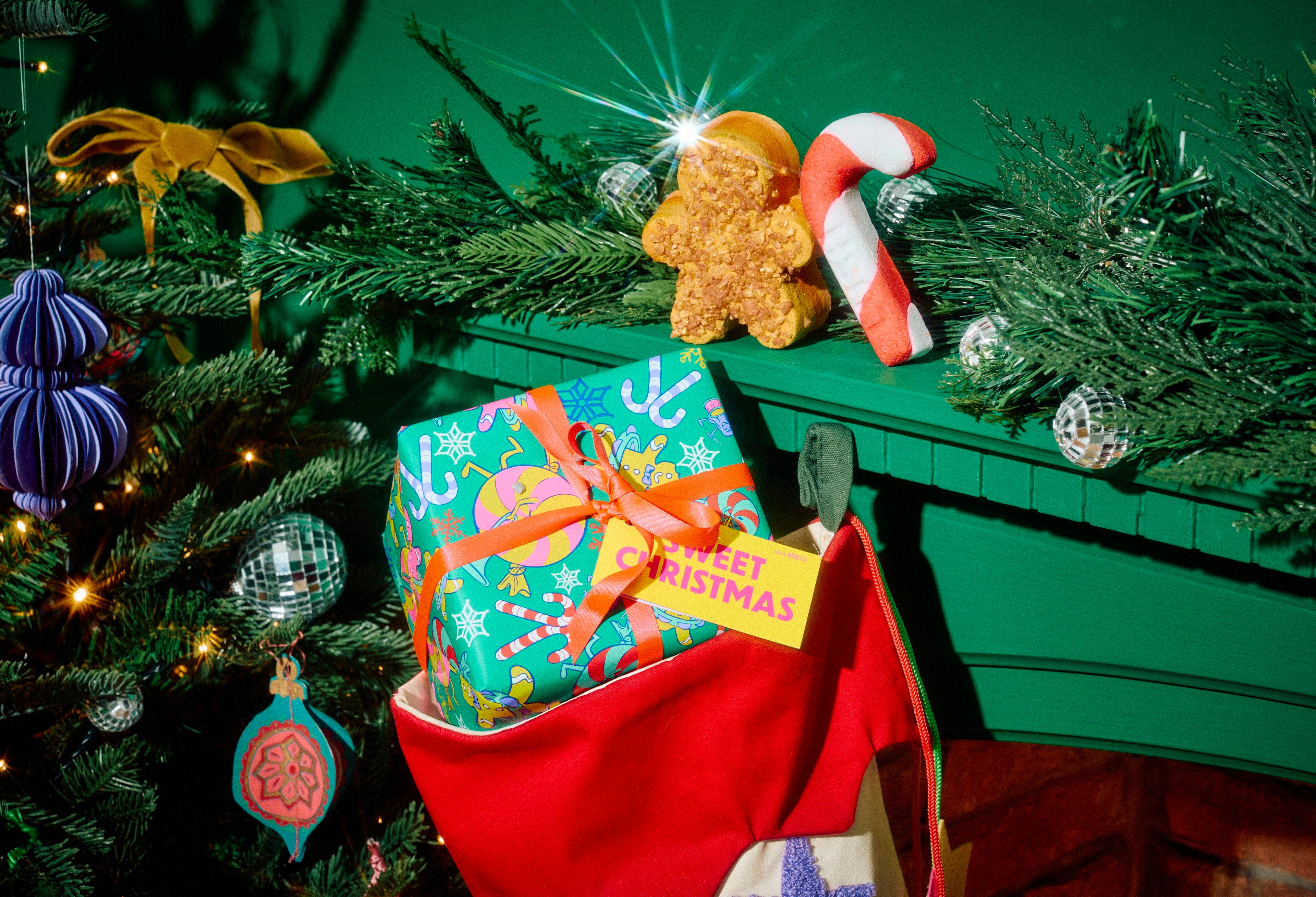 A green festive-patterned Lush 'Sweet Christmas' gift box with a yellow tag tucked inside a red stocking, with a gingerbread product and candy cane bubble bar resting on a green mantelpiece alongside a decorated Christmas tree.
