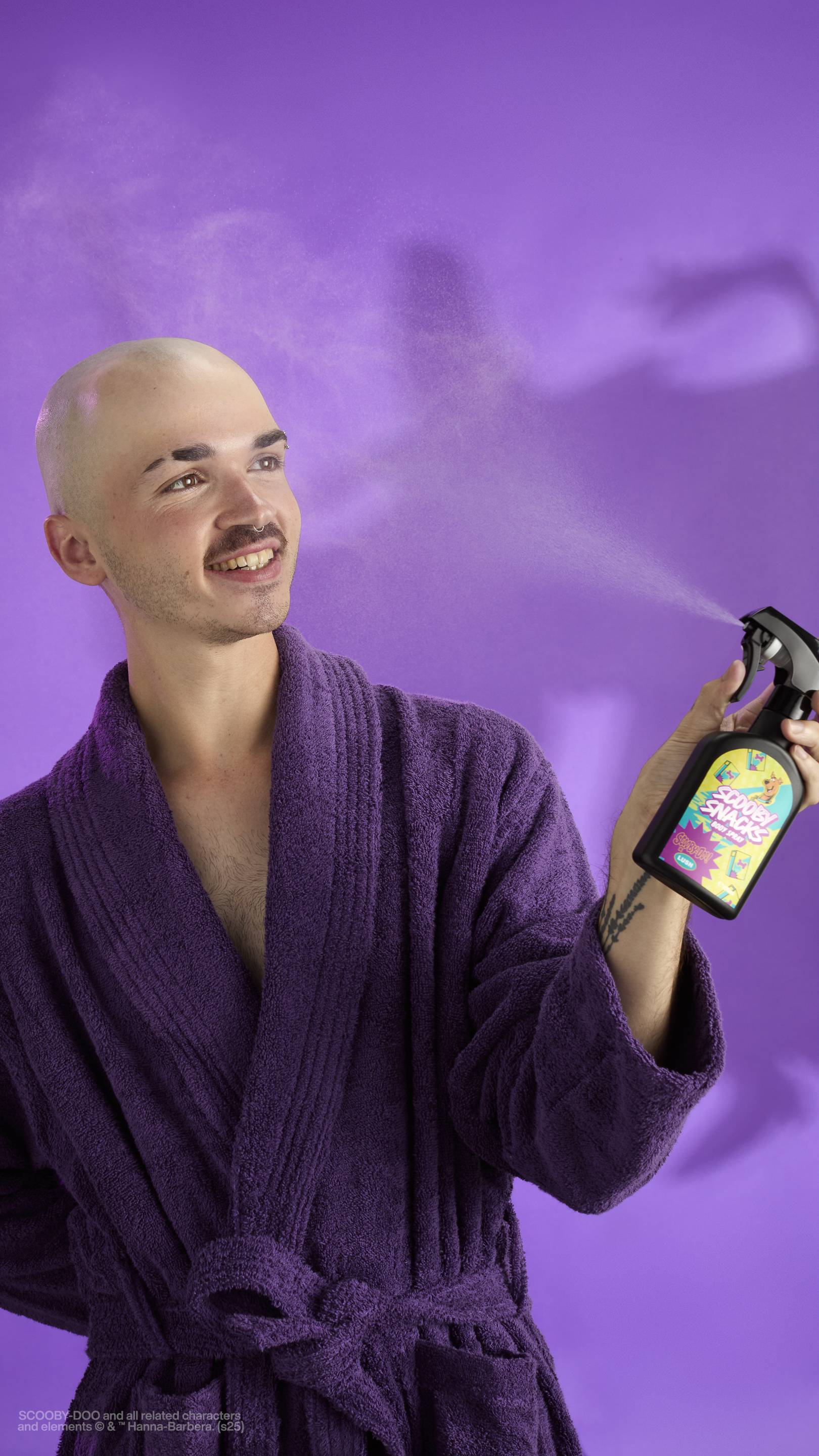 A person with a shaved head and a moustache, wearing a purple bathrobe, smiles as they spray a Lush Scooby Snacks body spray bottle. A fine mist is visible coming out of the nozzle. The background is a solid purple with an eerie shadow.