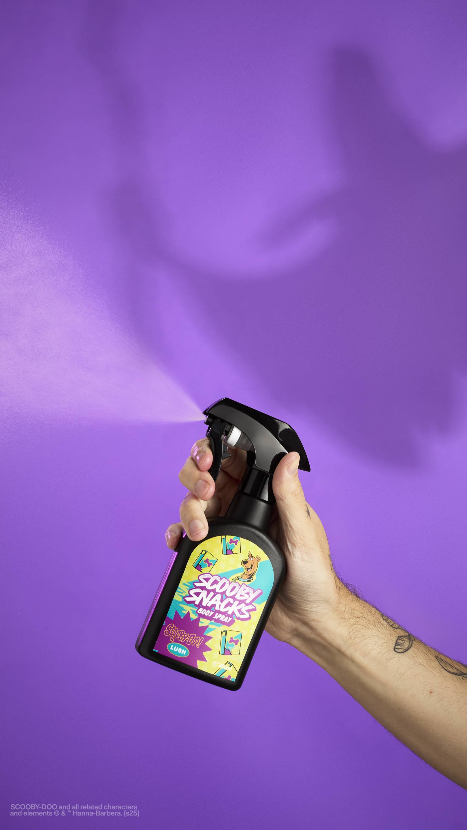The image shows a hand holding the Lush Scooby Snacks body spray bottle with a colourful label. They are spraying the product toward the left side of the frame, and a fine mist is visible. A purple background shows a large, eerie shadow.