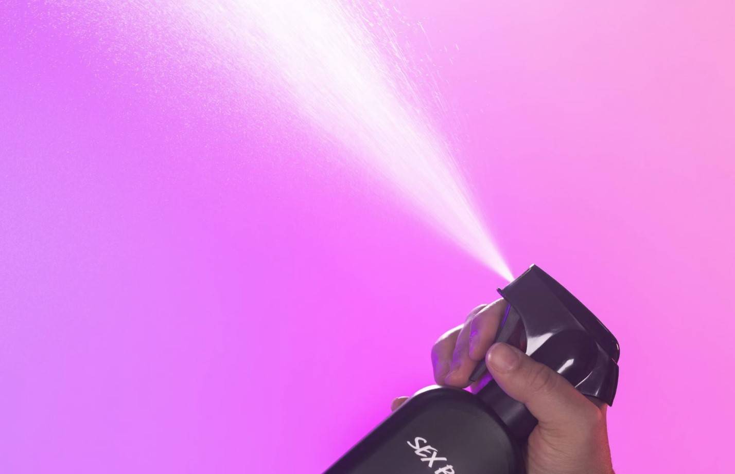Sex Bomb body spray is being held and spritzed into the air on a purple and pink background.