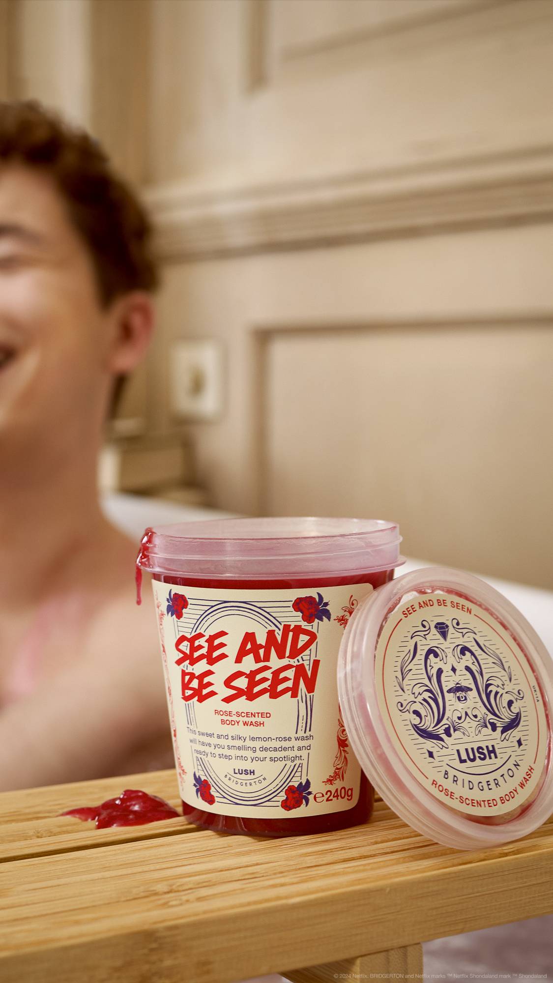 The image shows an out-of-focus model bathing in the background. The foreground focuses on the open, clear, LUSH pot of See And Be Seen body wash sitting on a wooden bath tray. Some product has been spilled. 