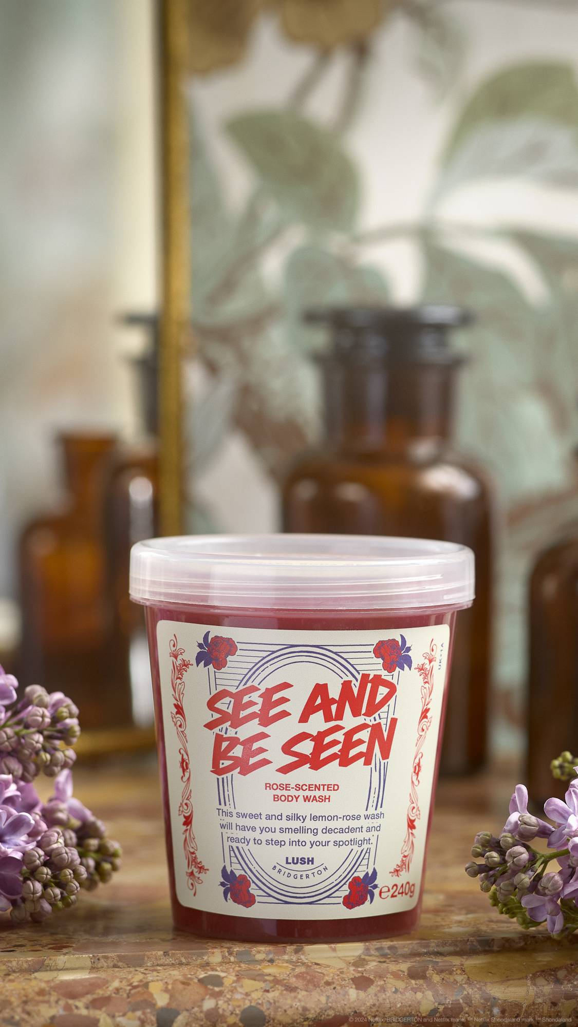 An out-of-focus background of leaves and what appears to be a dressing table. The focus is the clear LUSH pot of See And Be Seen body wash placed on a marbled counter with flowers on either side.