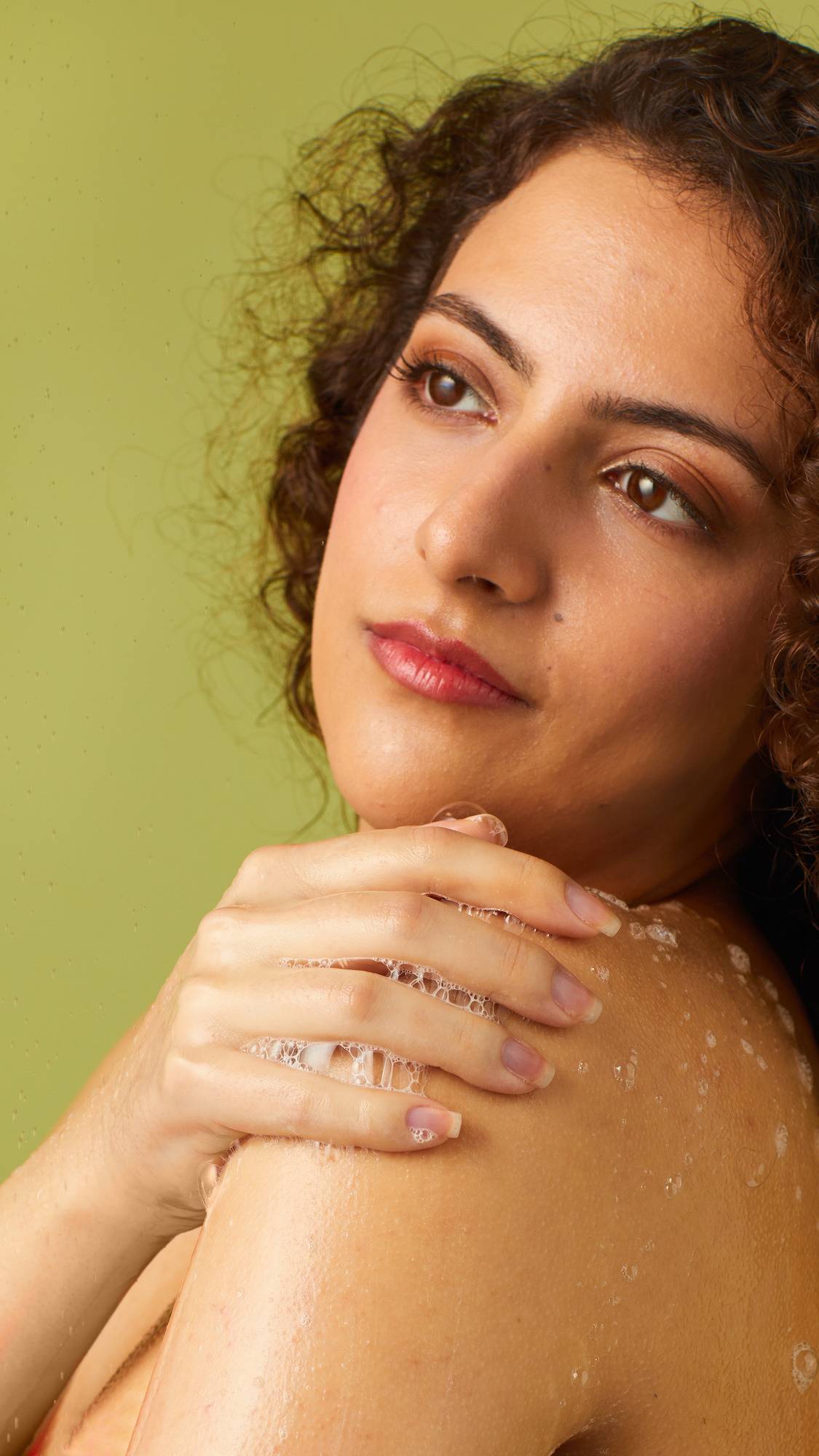 A close-up captures a person with curly dark hair washing in the shower against a light green background. Water droplets fall around them as they rest a soapy hand on their bare shoulder, lathering the product onto their skin.