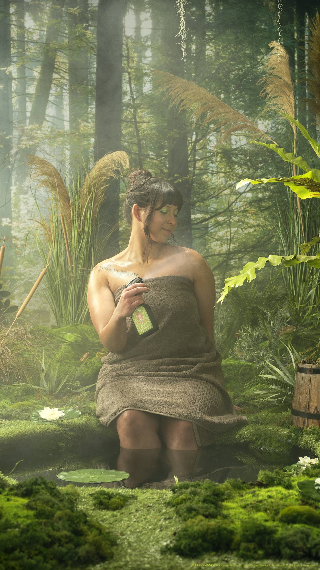 The model is sitting, wrapped in a fluffy brown towel, by the edge of the pond with their legs dipped in. They are surrounded by moss and shrubbery as they use the body spray. 