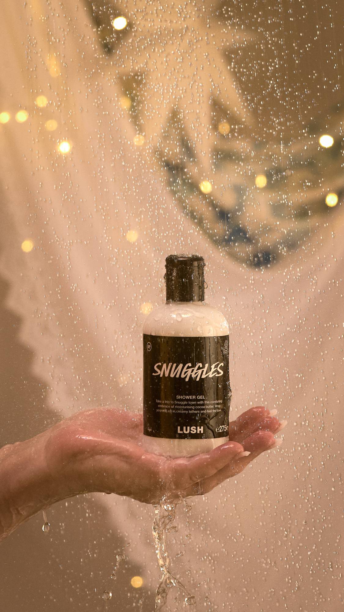 A close-up of the model's hand holding the Snuggles shower gel bottle under running shower water. 