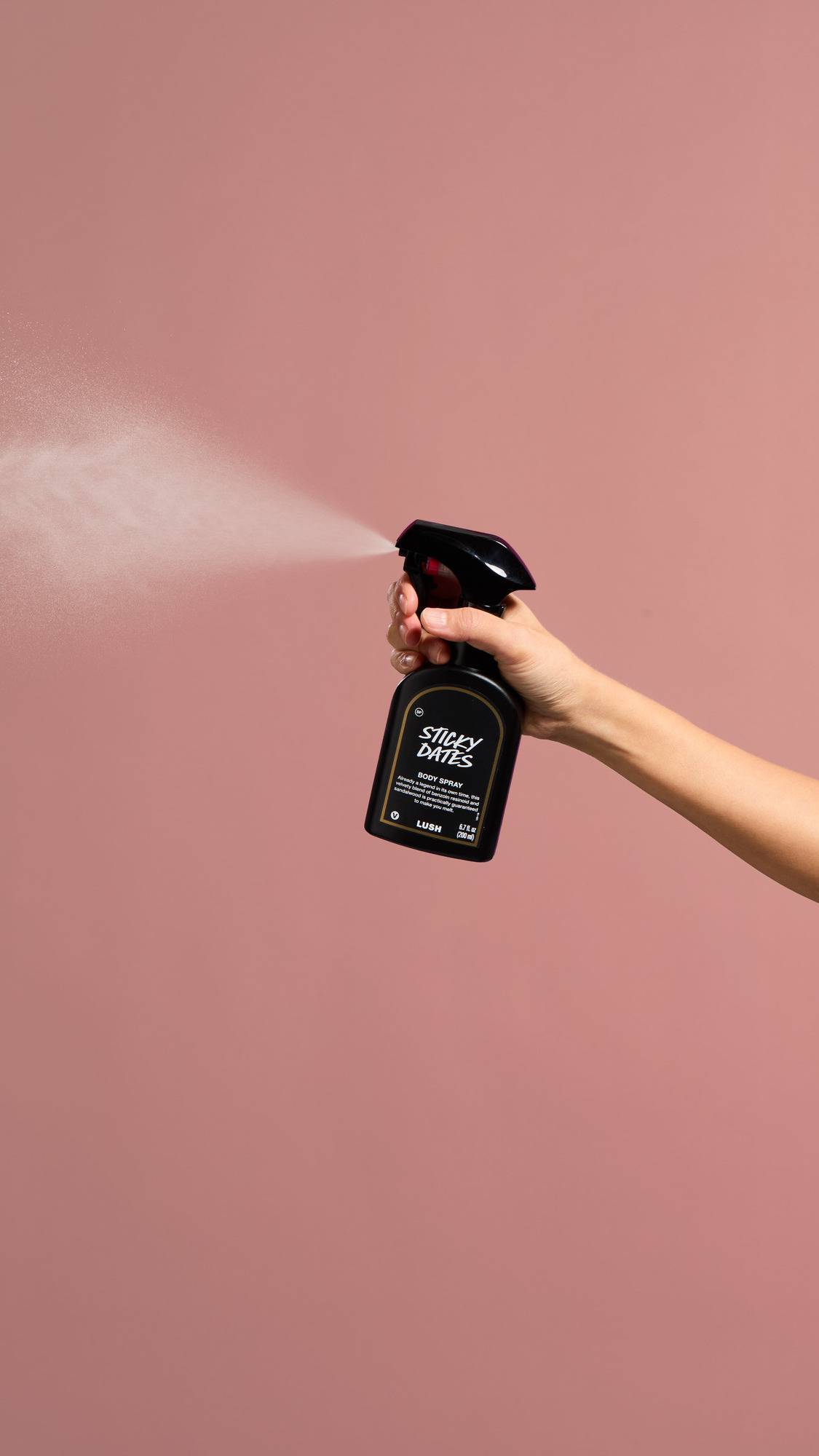 A hand holds a black bottle of "Sticky Dates" Body Spray, releasing a wide, fine mist into the air against a dusty pink background.