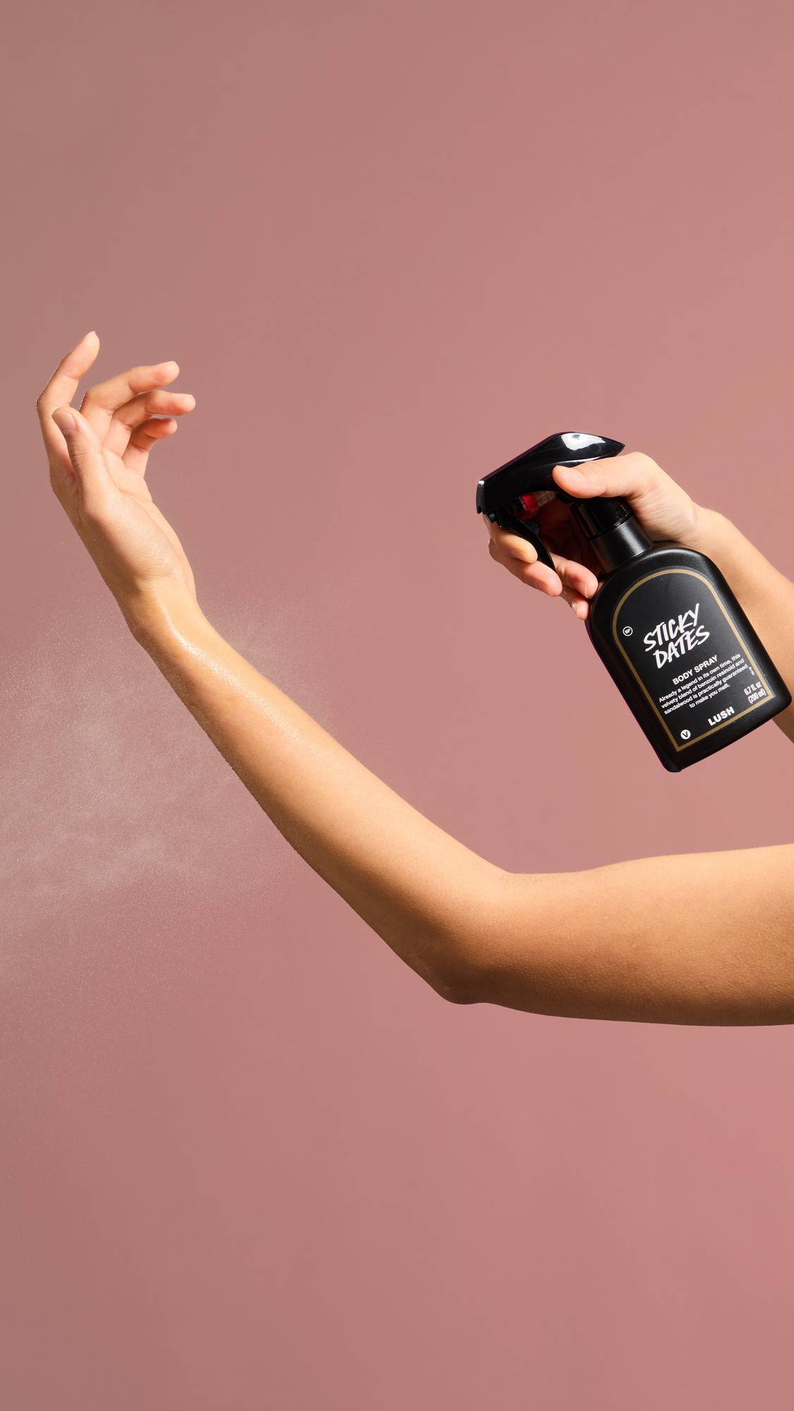 A person sprays a fine mist of "Sticky Dates" Body Spray onto their outstretched forearm against a dusty pink background. The black spray bottle features a label describing the scent as a blend of "benzoin resinoid and sandalwood".