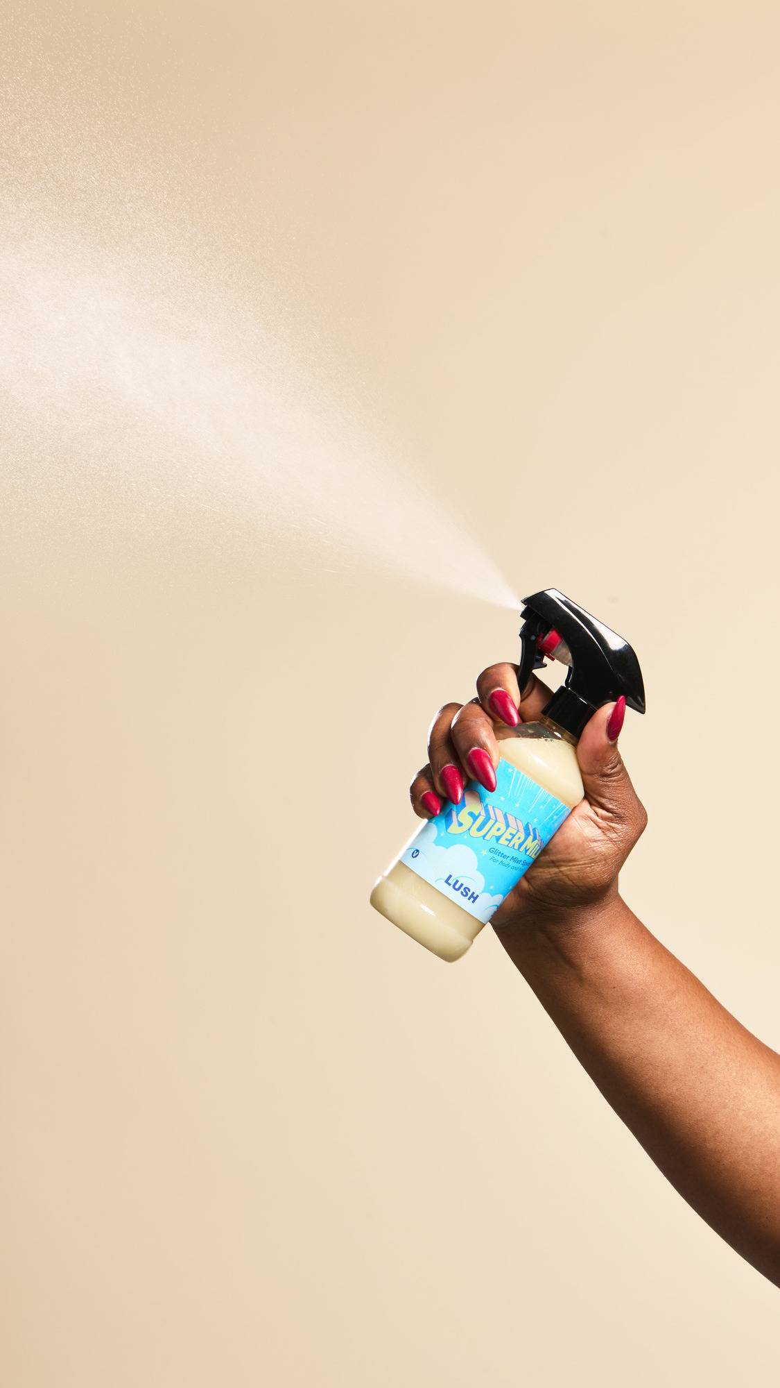 A hand with long red nails sprays a fine mist from a bottle of "Super Milk Glitter Mist" against a beige background. The clear bottle reveals a pale yellow liquid and features a bright blue label decorated with clouds and stars.