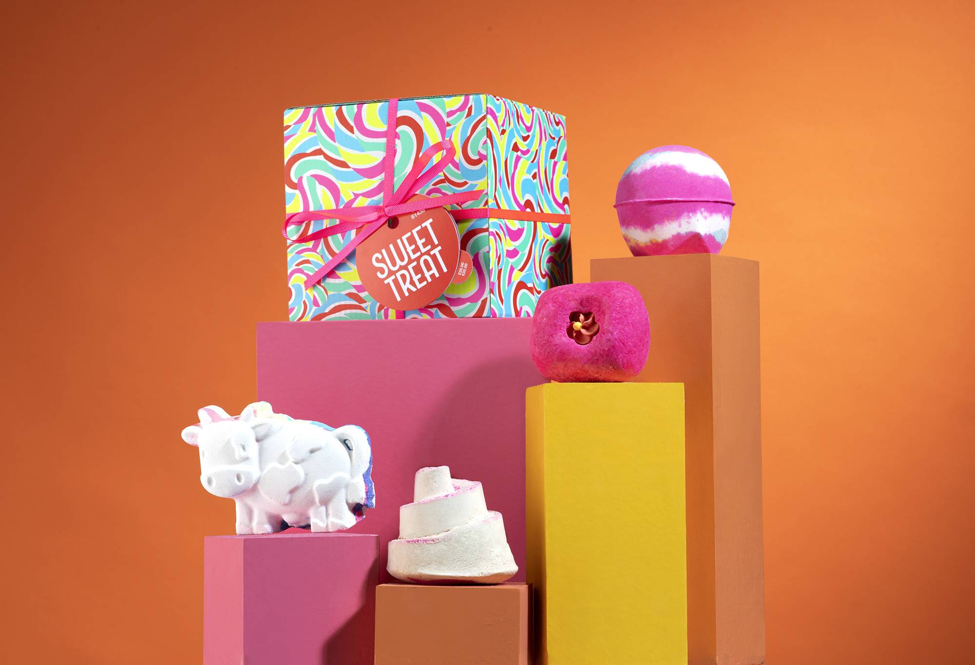 Sweet Treat gift, in front of an orange background, surrounded by its products on pink, orange and yellow plinths.