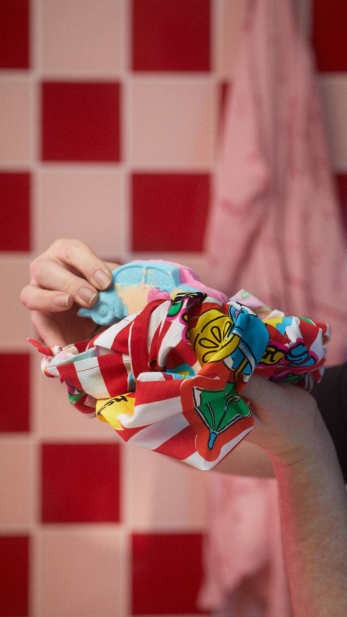 The image shows a close-up of the model holding the Lush bath bomb half-wrapped in the bold, bright Sweets For My Sweet knot wrap.