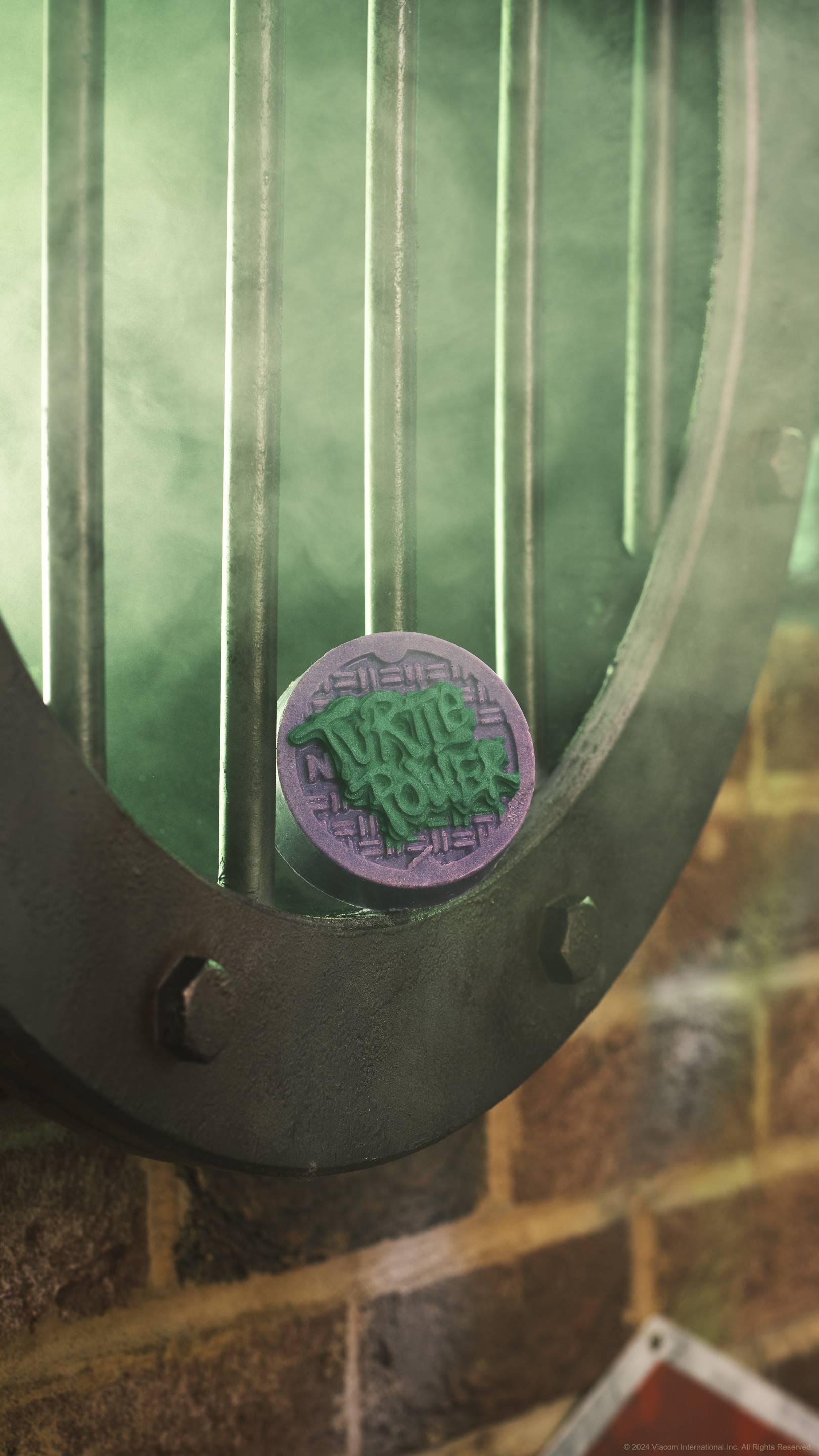 The image shows the close-up of a large side-facing metal pipe grate shrouded in green-tinted smoke. The Turtle Power solid body scrub is sat on the end of the pipe grate in central focus. 