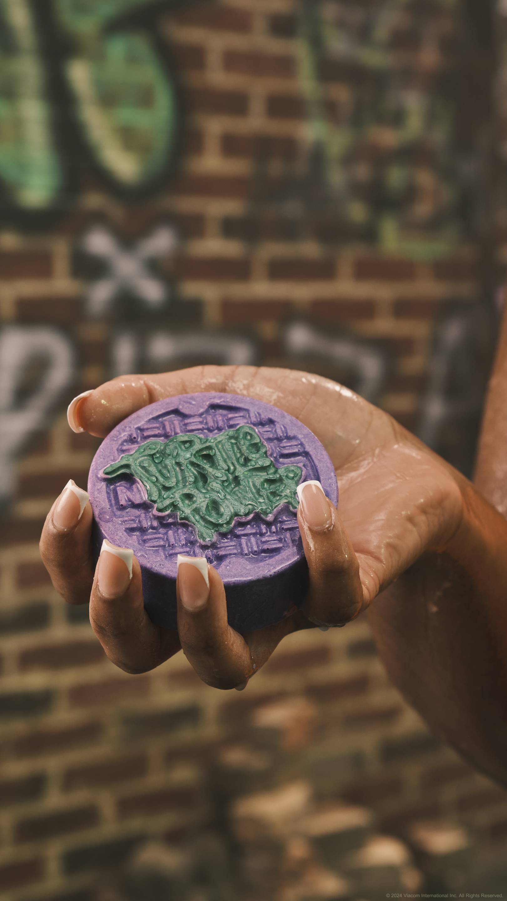 A close-up image of a model's hand with manicured nails holding the Turtle Power solid body scrub to the camera. The background is a blurred graffitied brick wall. 