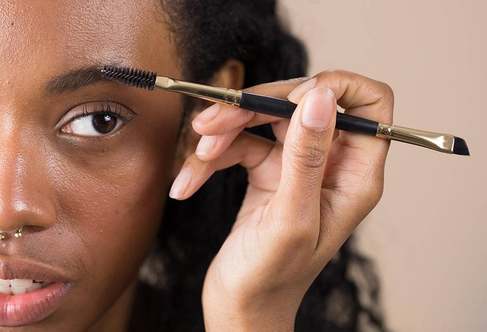 A person using U-N-I-Brow, a double ended brow brush, with a spoolie and an angled brush, to comb and style their brows.