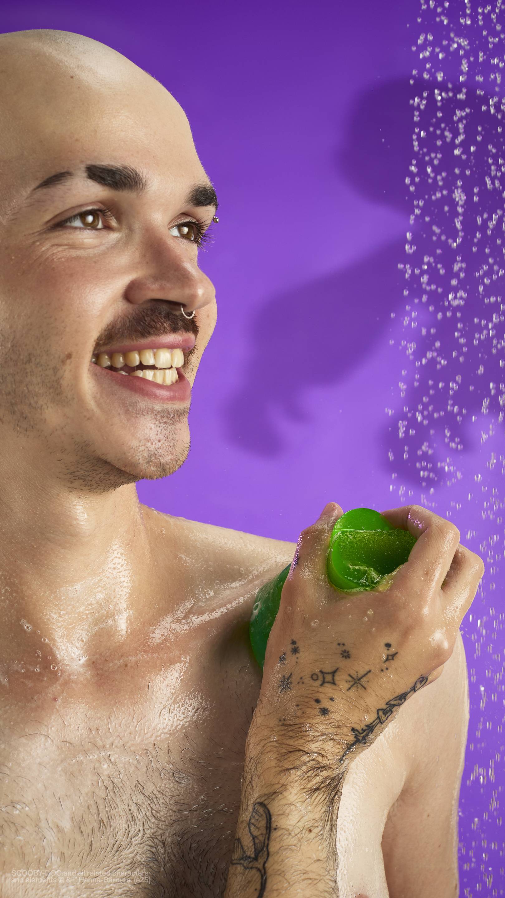 The image shows a solid purple background with out-of-focus falling shower water. A light-toned model with a shaved head and moustache is holding the Zoinks! shower jelly and creating a lather of suds on their shoulder. 