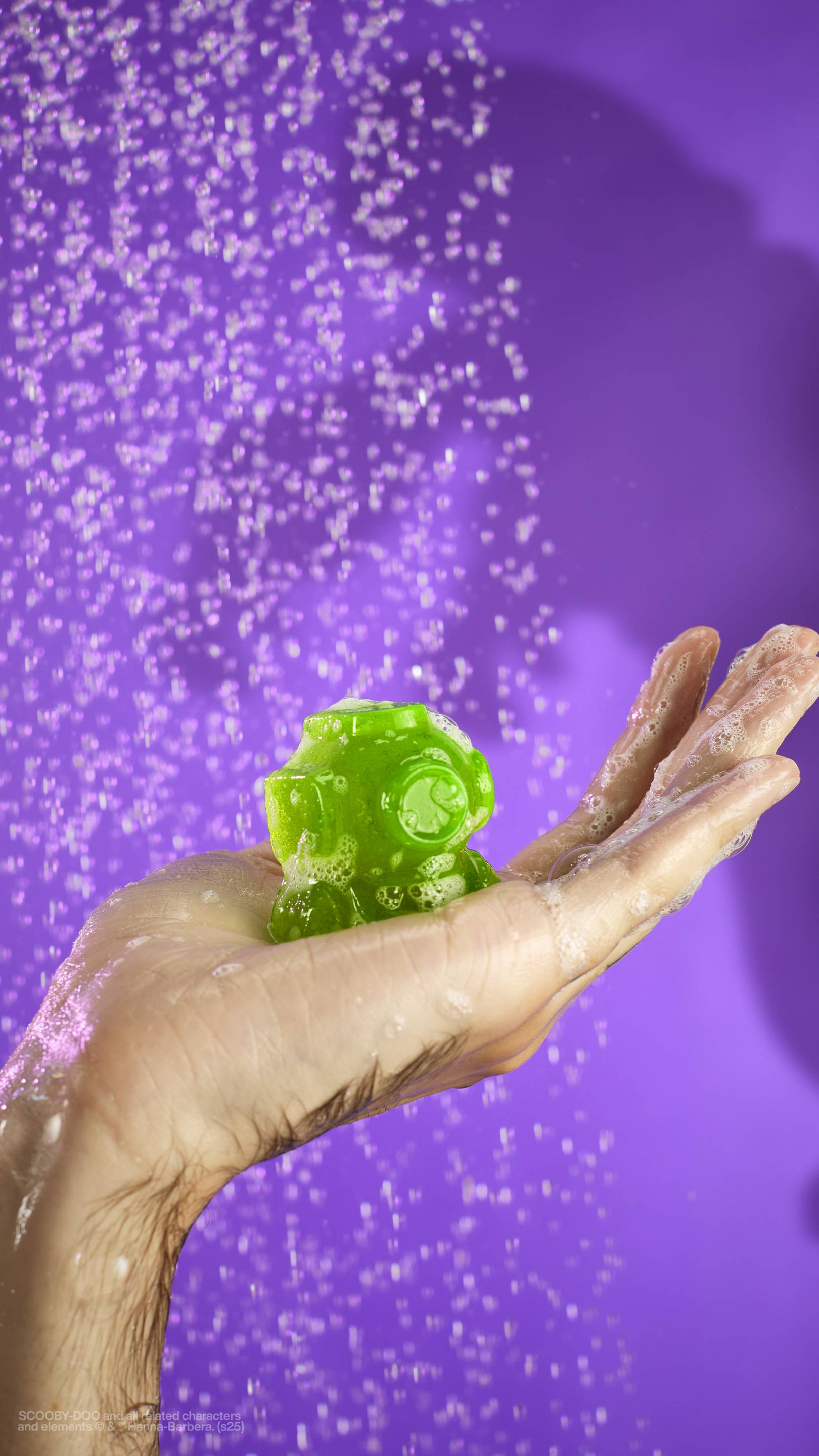 The image shows a solid purple background with out-of-focus falling shower water. A light-toned hand holds the embedded, classic scuba diver helmet-shaped shower gummy in their palm, coated with suds. 