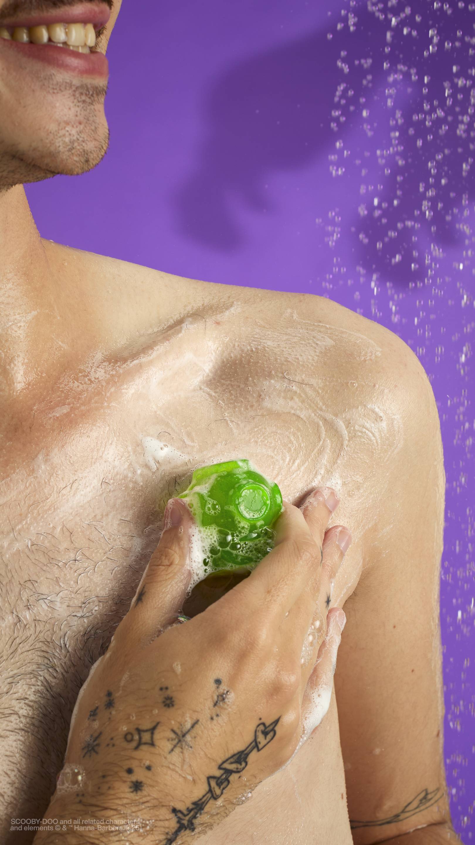 The image shows a solid purple background with out-of-focus falling shower water. A light-toned model is holding the embedded scuba diver helmed-shaped gummy from the Zoinks! shower jelly and creating a lather of suds on their shoulder. 