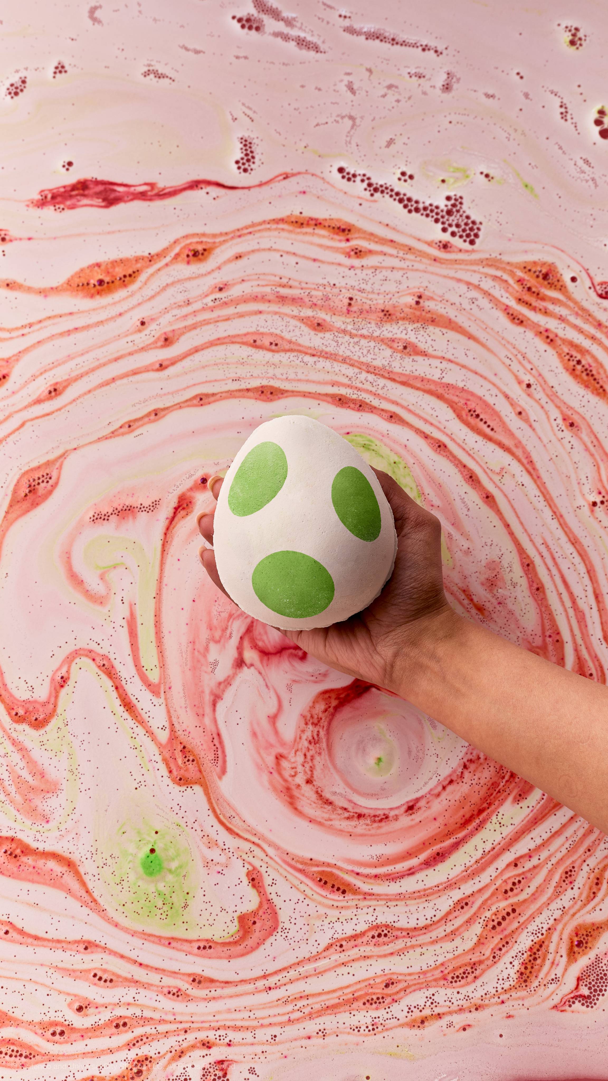 A hand holds a white Yoshi egg bath bomb with green spots over pink bathwater filled with red and light green foam swirls.