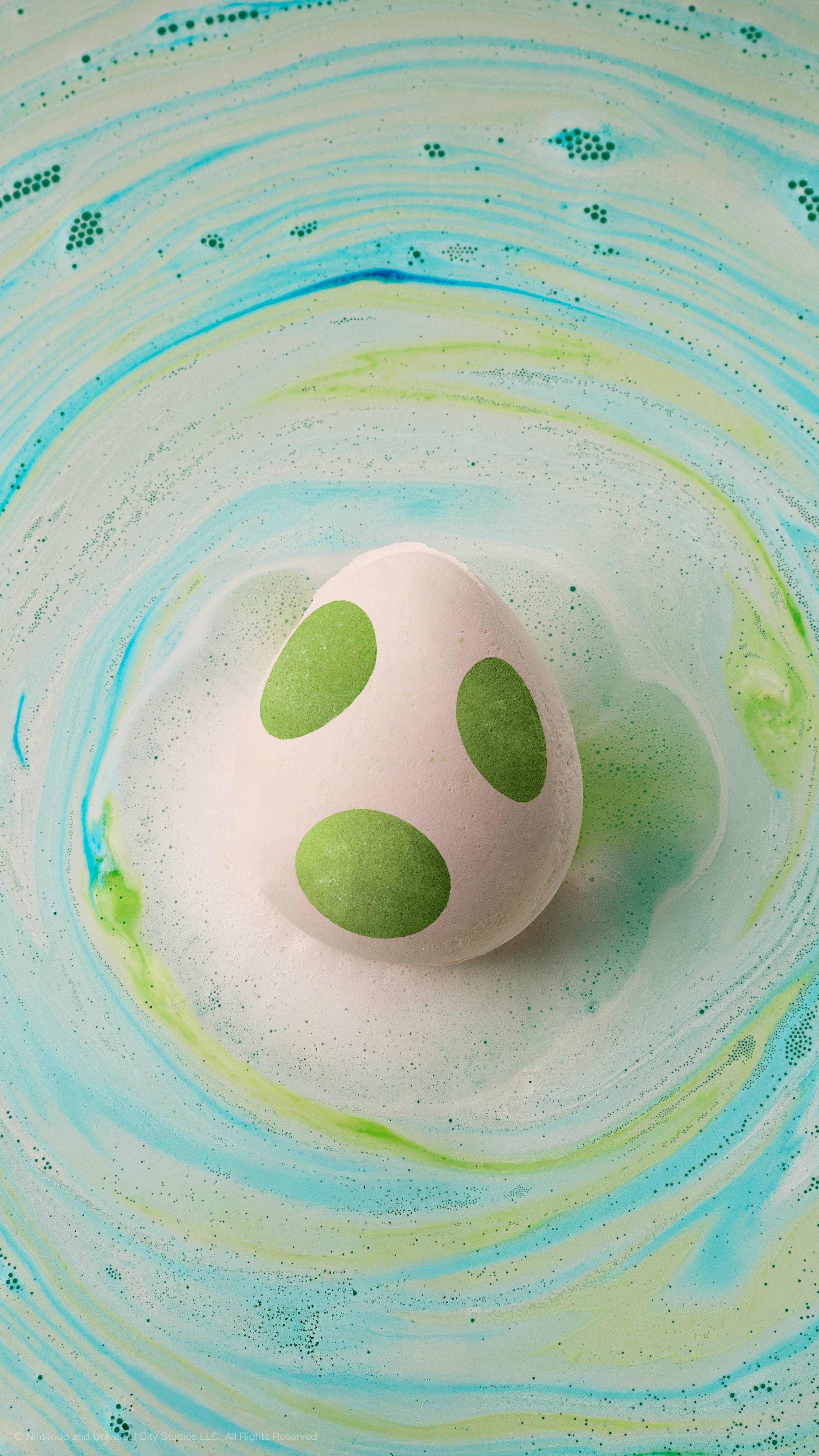 A white Yoshi egg bath bomb with green spots floats in water, surrounded by swirling patterns of green, blue, and white foam.