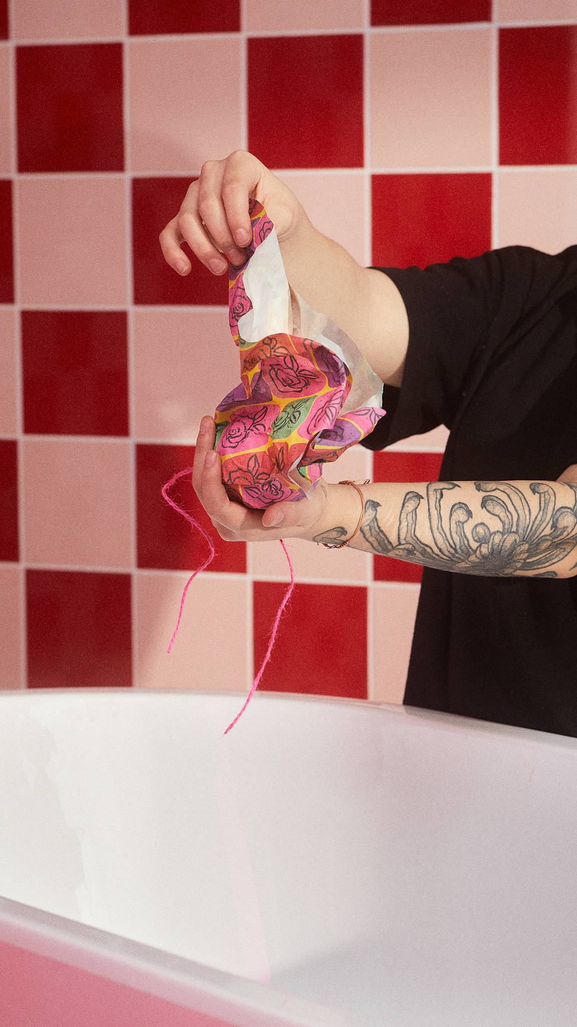 The image shows the model unwrapping the Abstract Roses gift wrap from a Lush bath bomb on a tiled background.