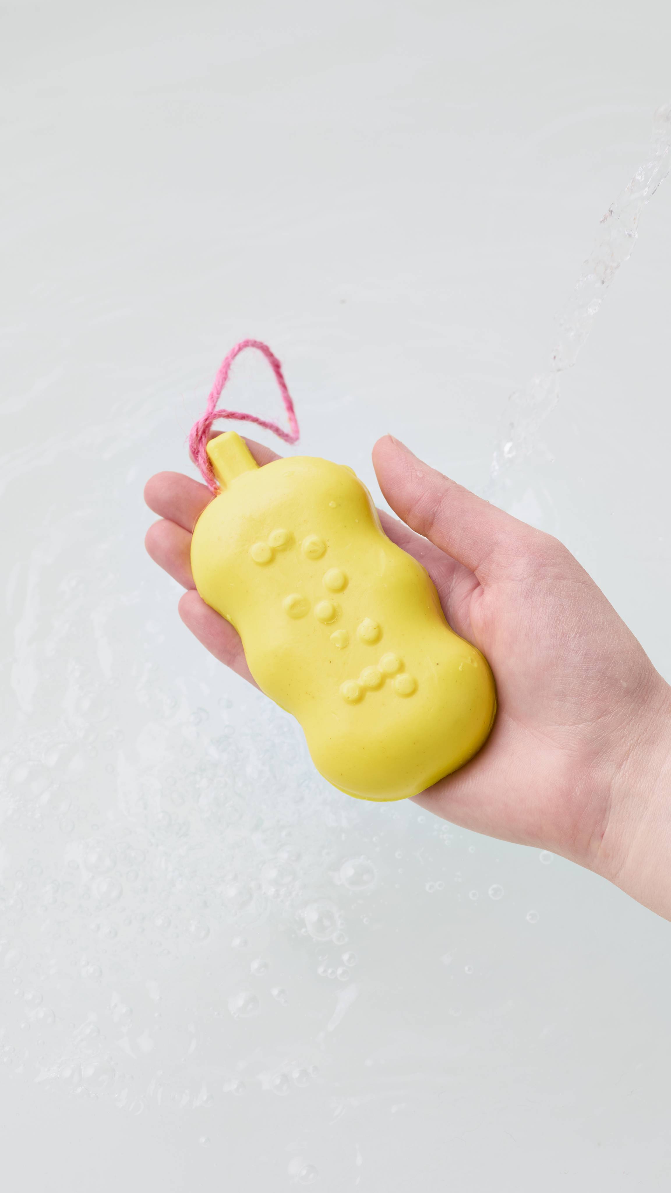 A hand holds a bright yellow soap bar with raised Braille lettering and a pink hanging cord, resting above rippling water as running water streams in.
