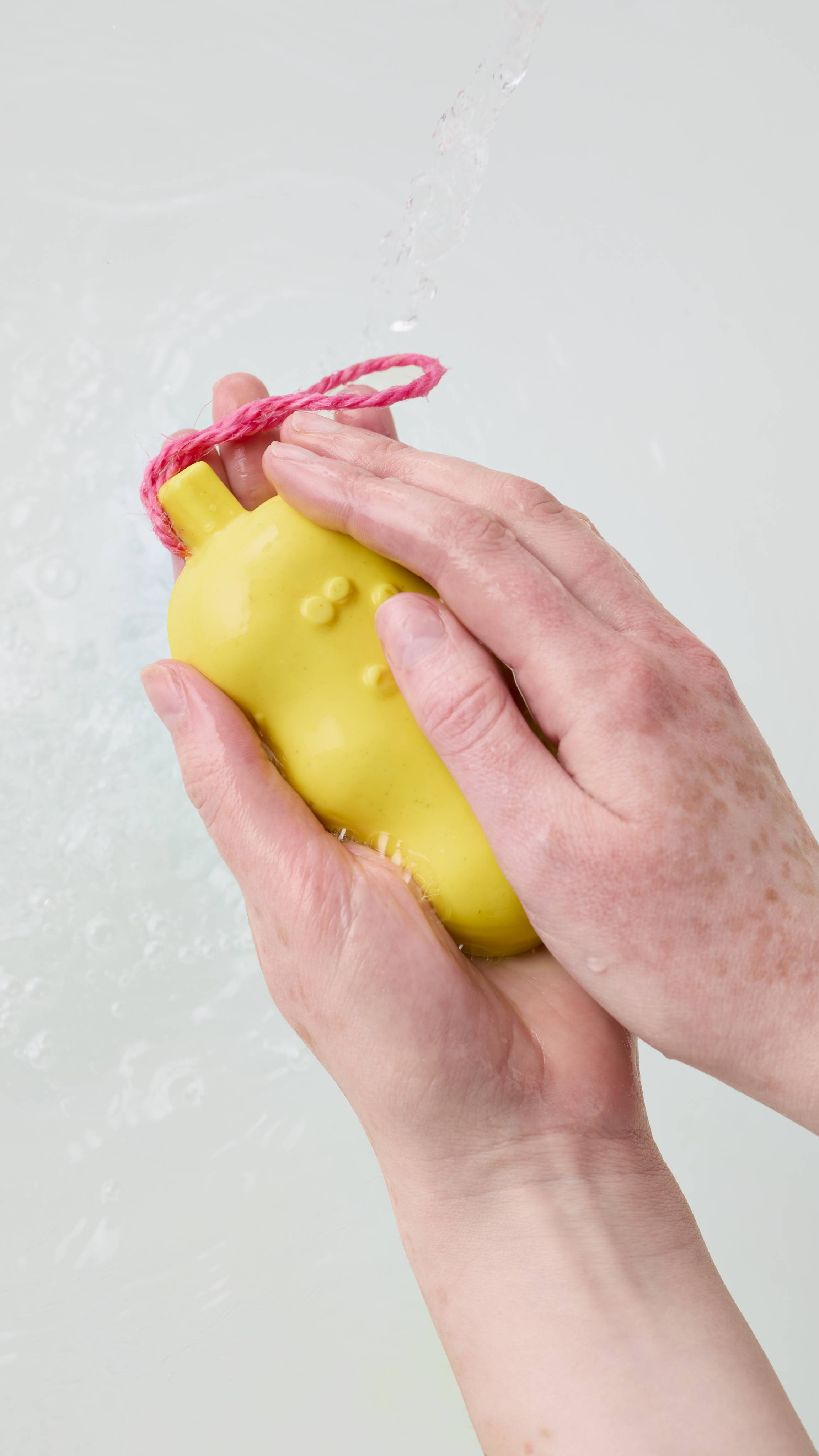 Two hands hold and gently lather a bright yellow soap bar with a pink hanging cord under a stream of running water, set against a bath of clear, rippling water.