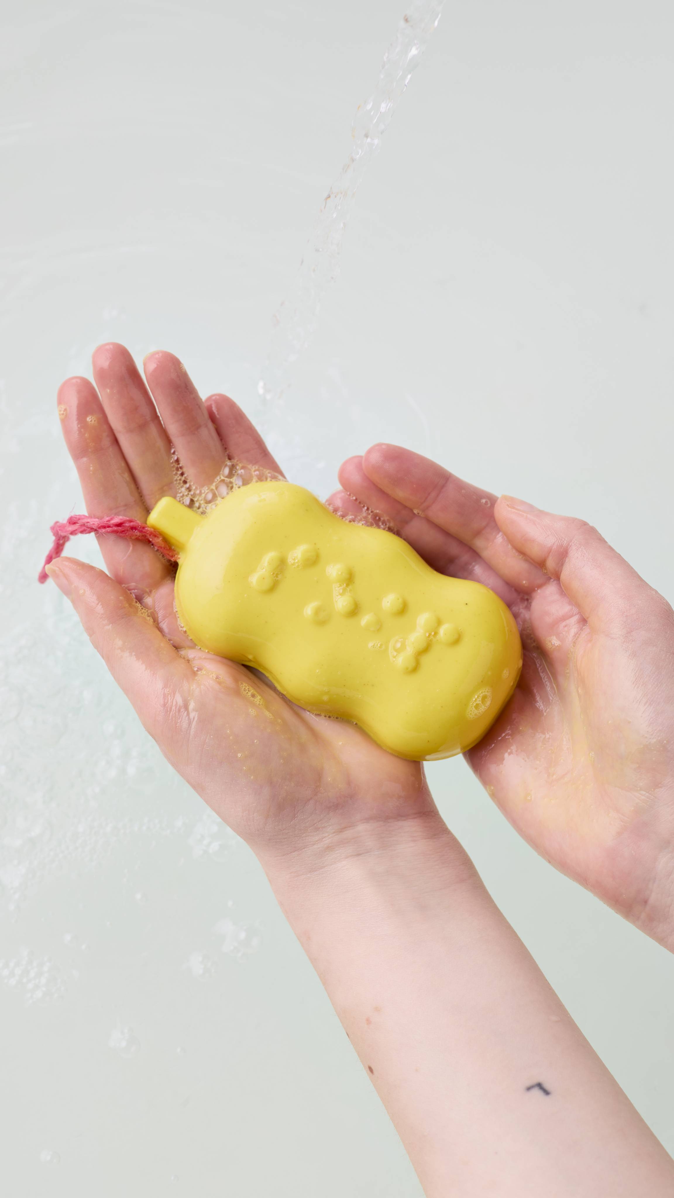 Two hands cup a bubbly, bright yellow soap bar with raised Braille details and a pink cord, held beneath a stream of running water over a bath filled with clear water.