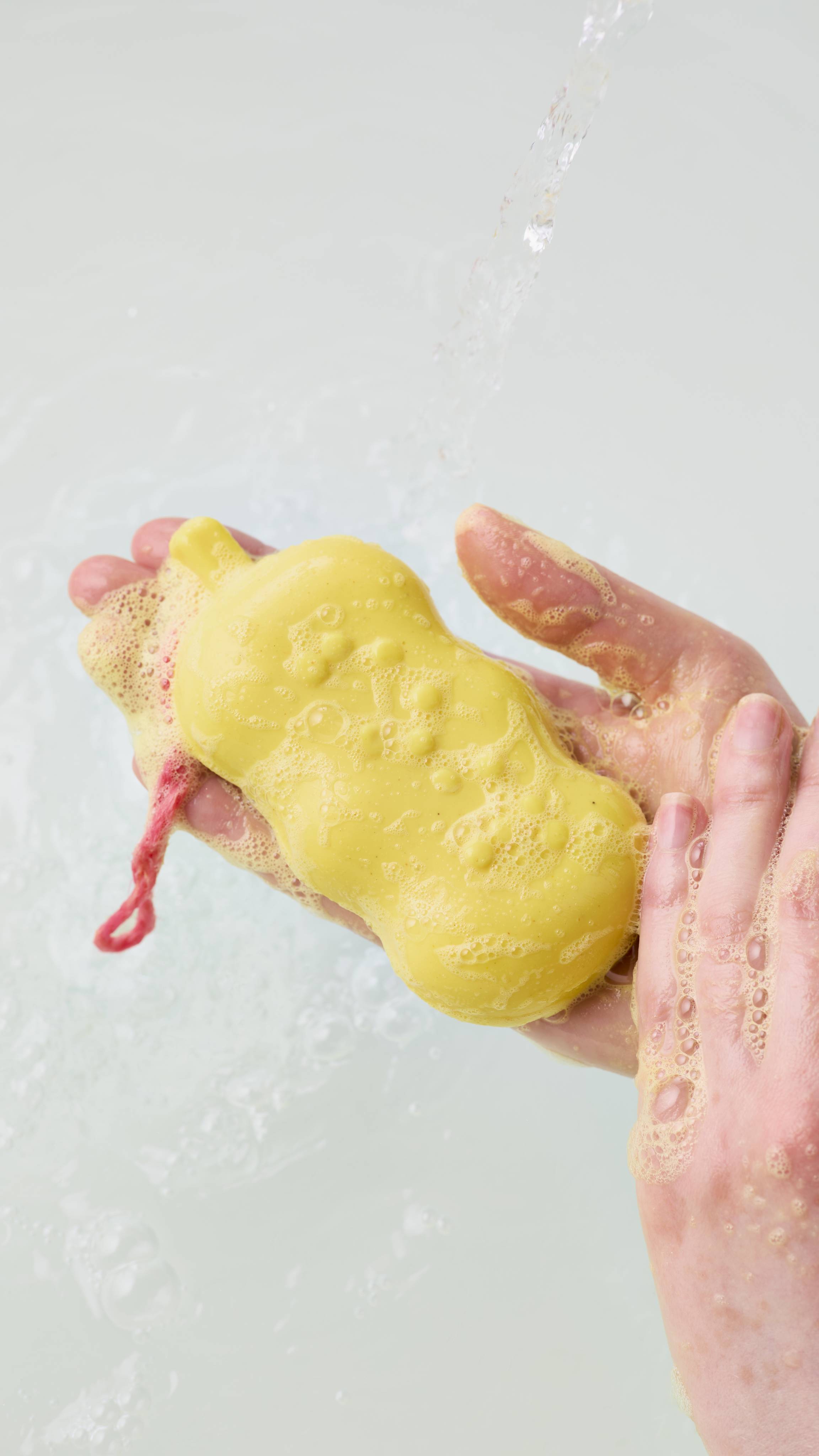 A yellow soap bar covered in rich, frothy bubbles rests in two wet hands under a stream of running bathwater, showcasing its raised Braille lettering and pink hanging cord.