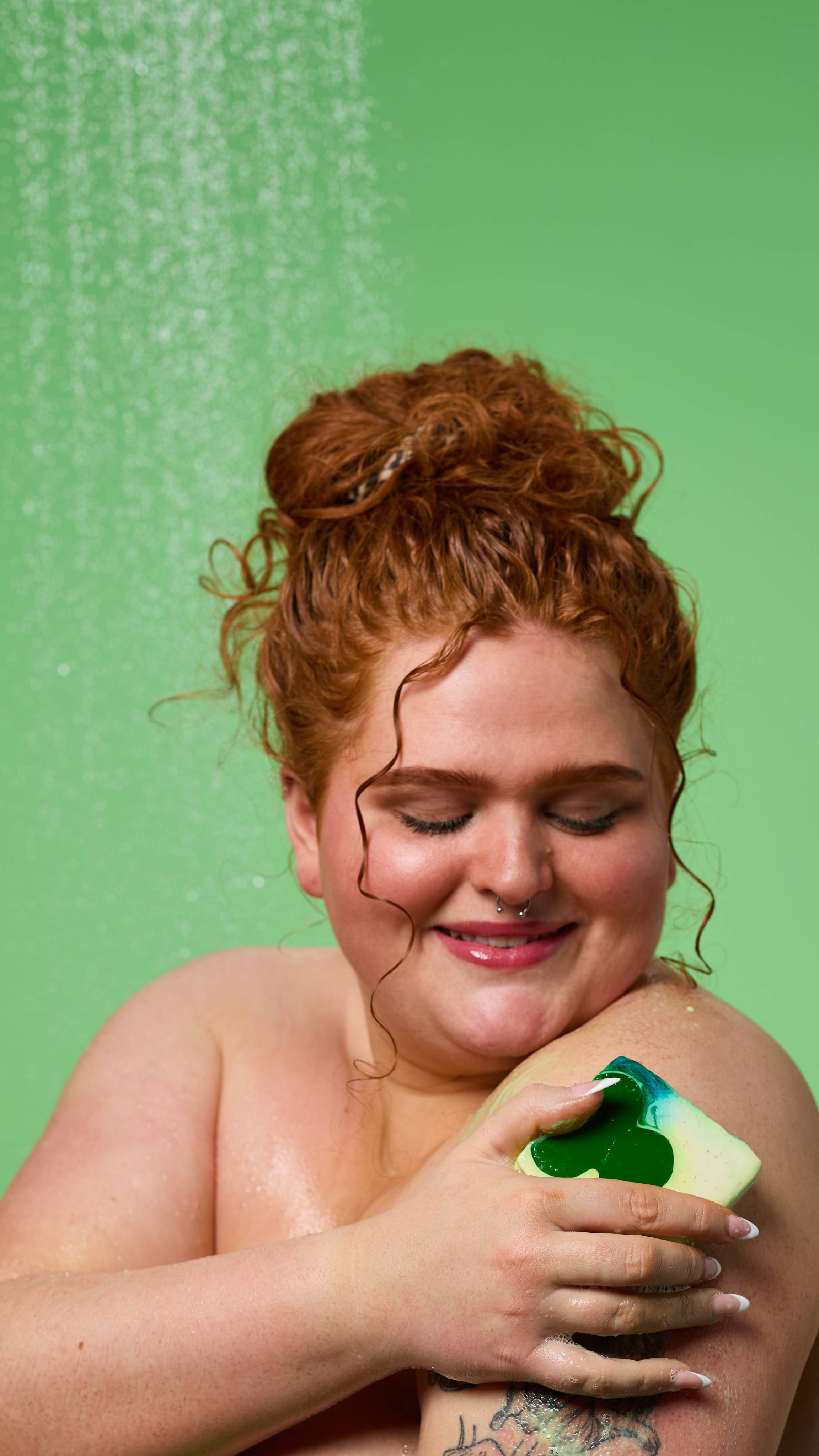 The model is standing under running shower water on a pale green background. Their curly hair is tied up as they lather their shoulder with the Alban Eilir soap. 