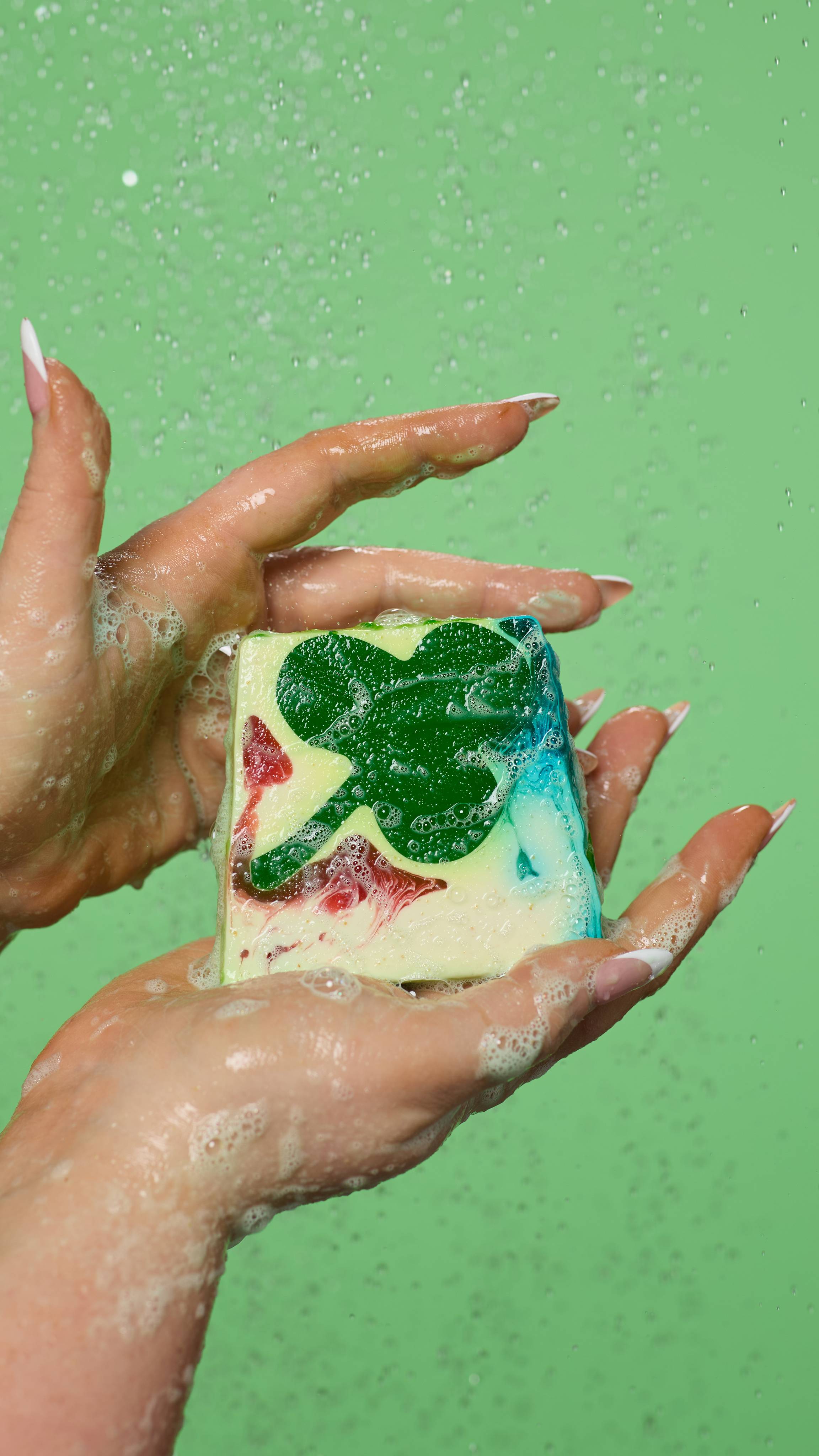 A close-up of the model's hands, lathered up with soapy suds while holding the Alban Eilir soap. Water is falling from above and there is a light green background.