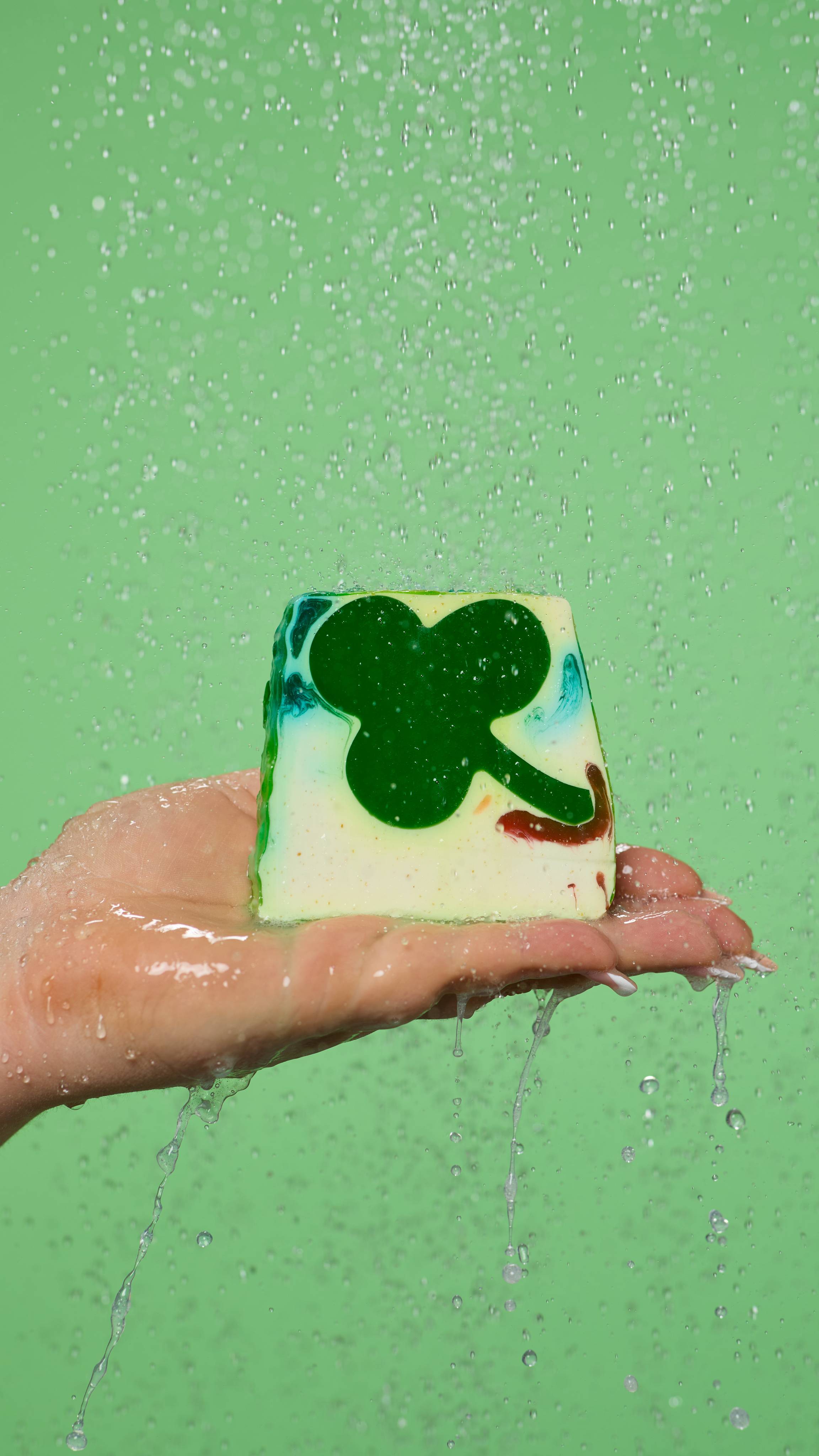 The image shows a close-up of the model's hand held flat under running shower water as the Alban Eilir soap is standing on their palm.