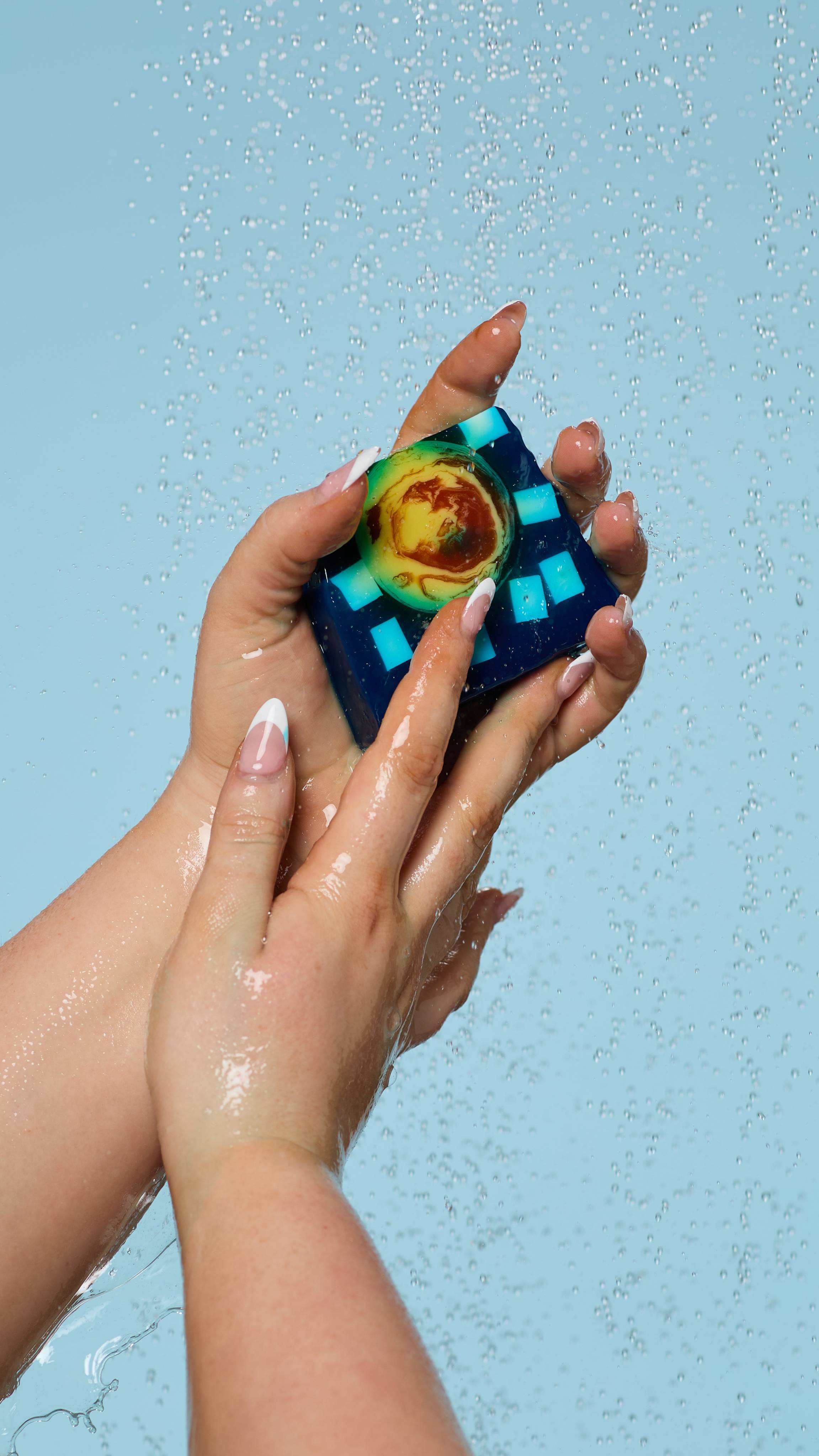 A close-up of the model's hands, lathered up with soapy suds while holding the Alban Arthan soap. Water is falling from above and there is a pale blue background.