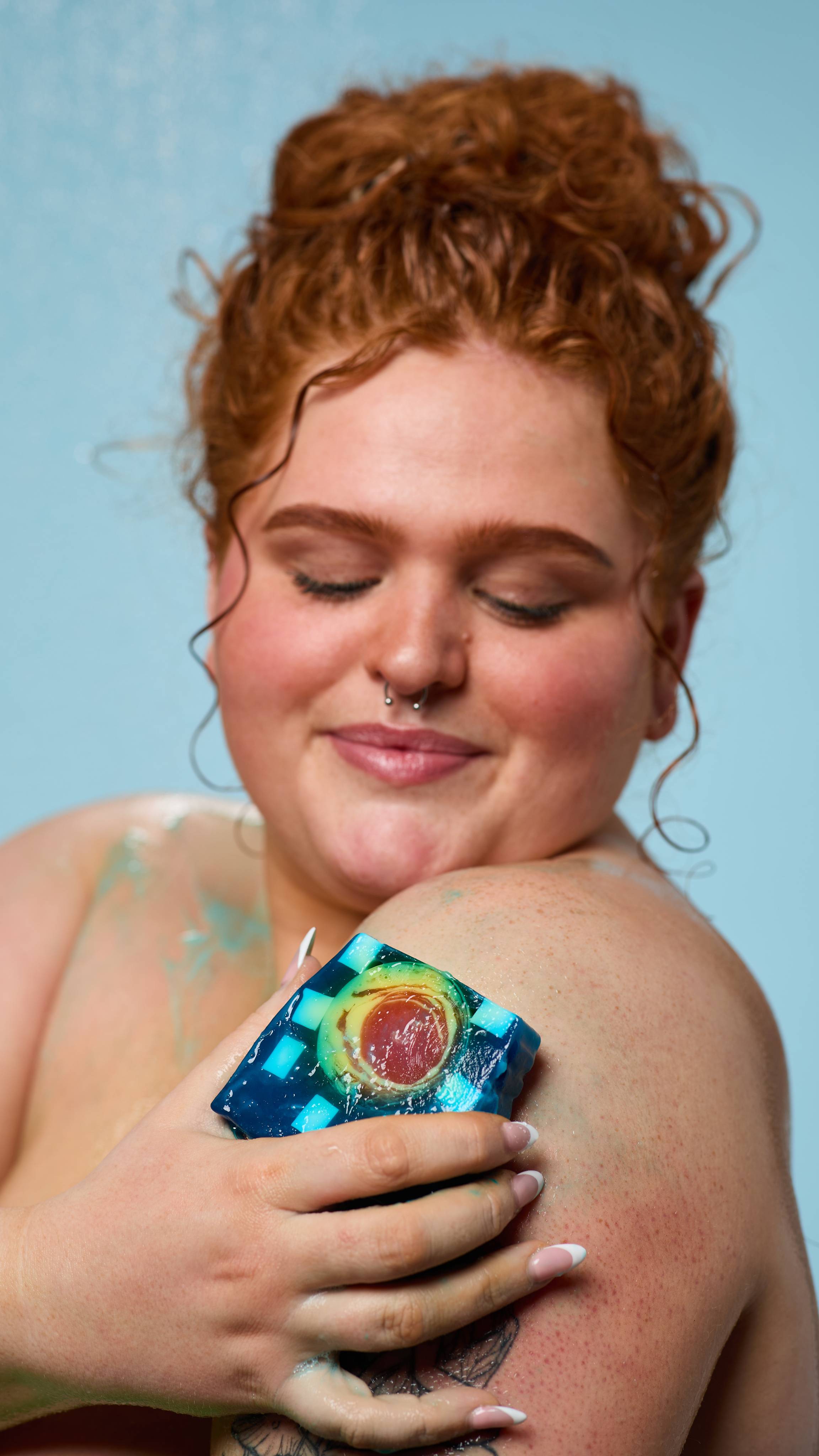 The model is standing under running shower water on a pale blue background. Their curly hair is tied up as they lather their shoulder with the Alban Arthan soap. 