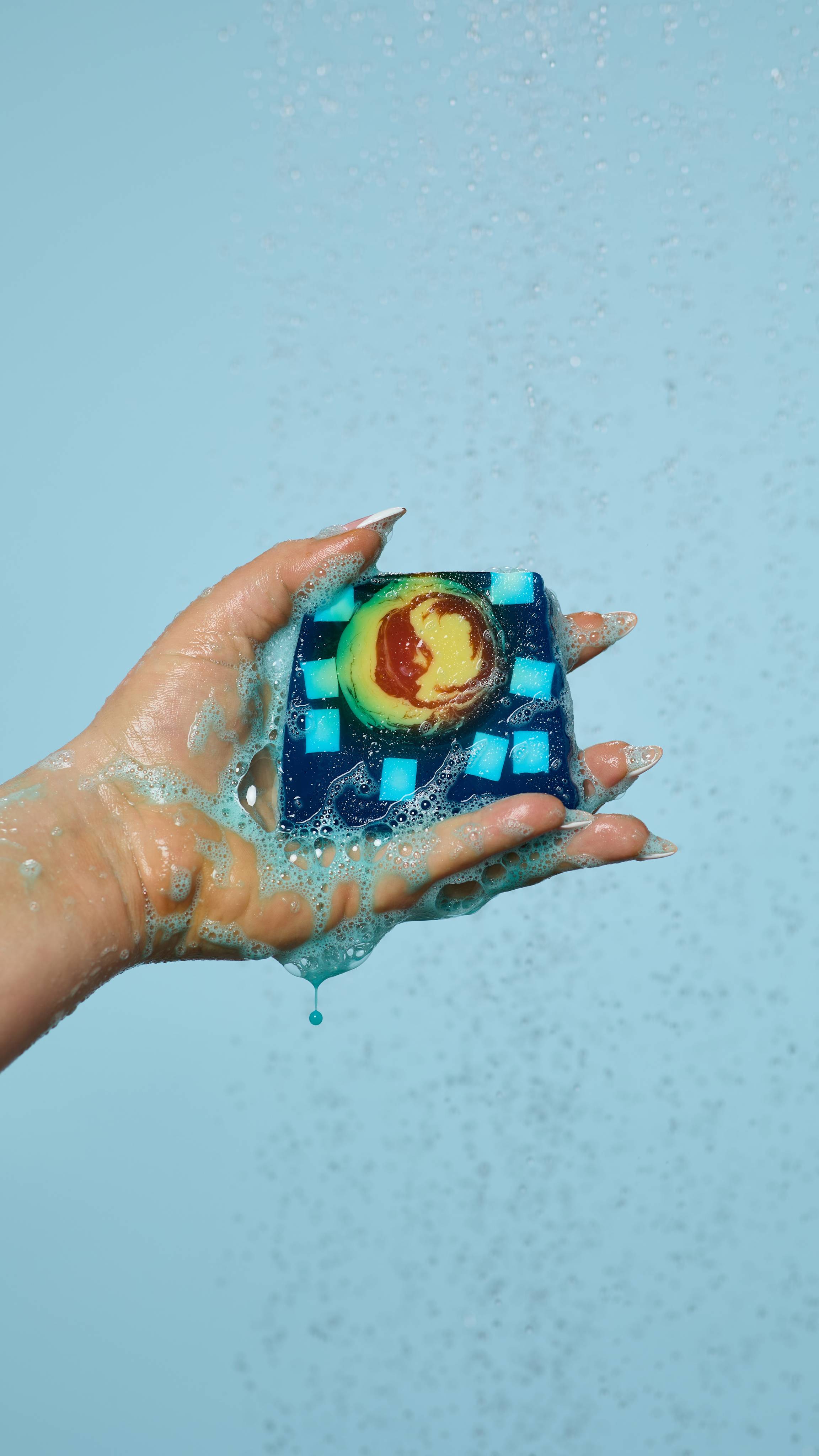 A close-up of the model's hand, lathered up with blue, soapy suds while holding the Alban Arthan soap. Water is falling from above and there is a pale blue background.