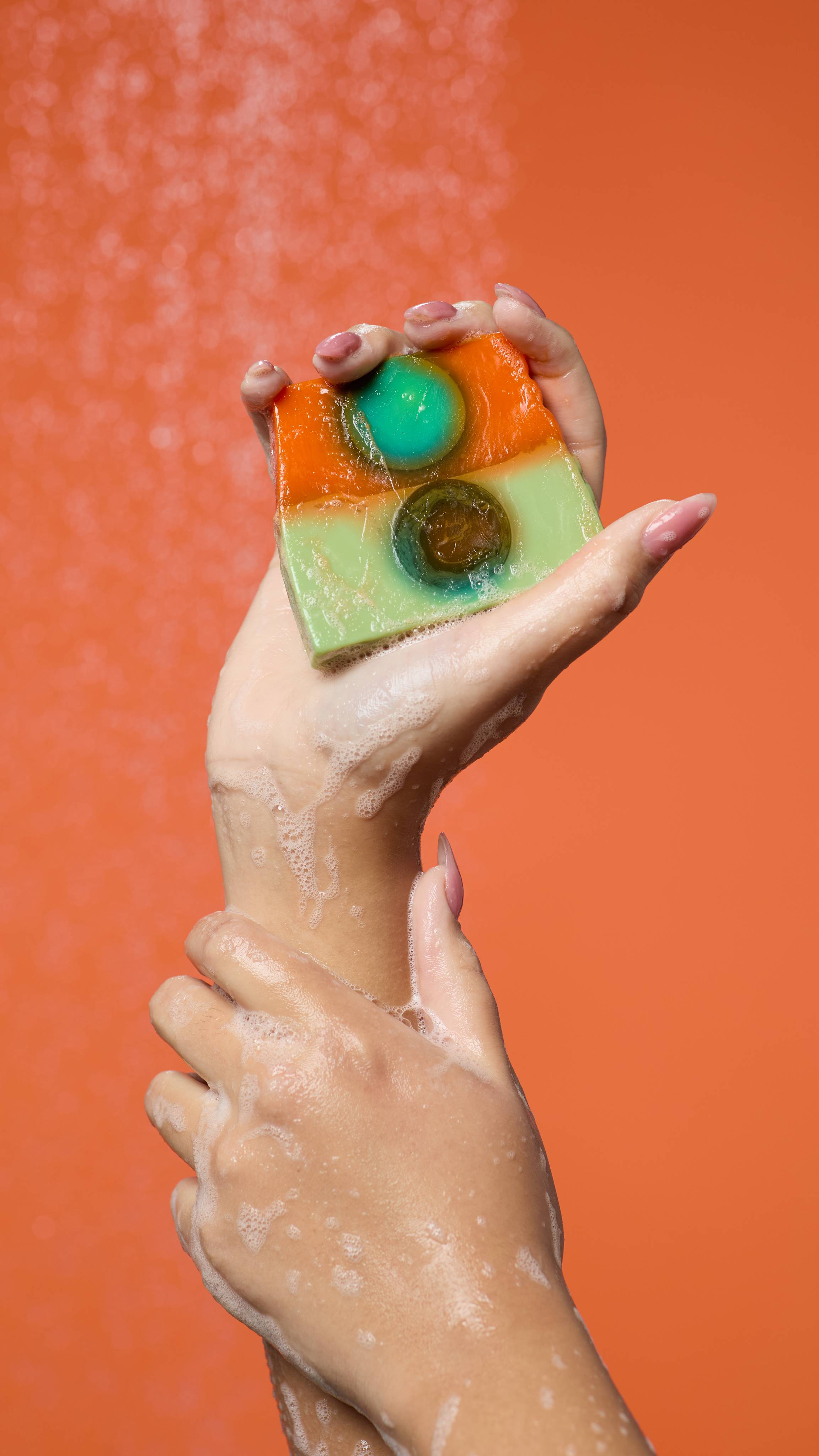 The image shows a blurred background with running shower water. The model's forearms can be seen as one hand holds the Alban Elfed soap, gently laced in suds. 
