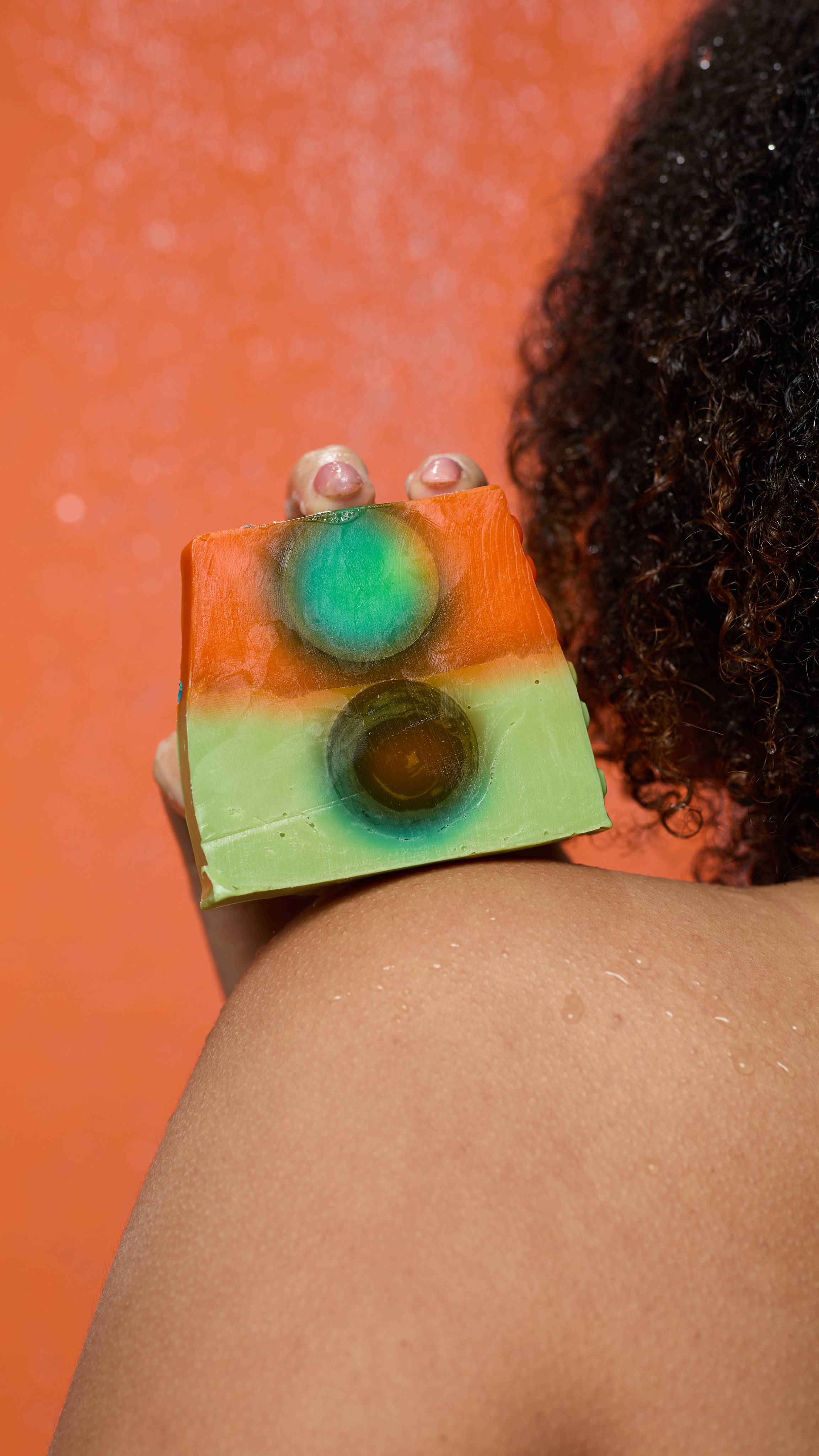 A close-up shot of the model's bare shoulder from behind, on a burnt-orange background. They are holding the Alban Elfed soap propped on their shoulder. 