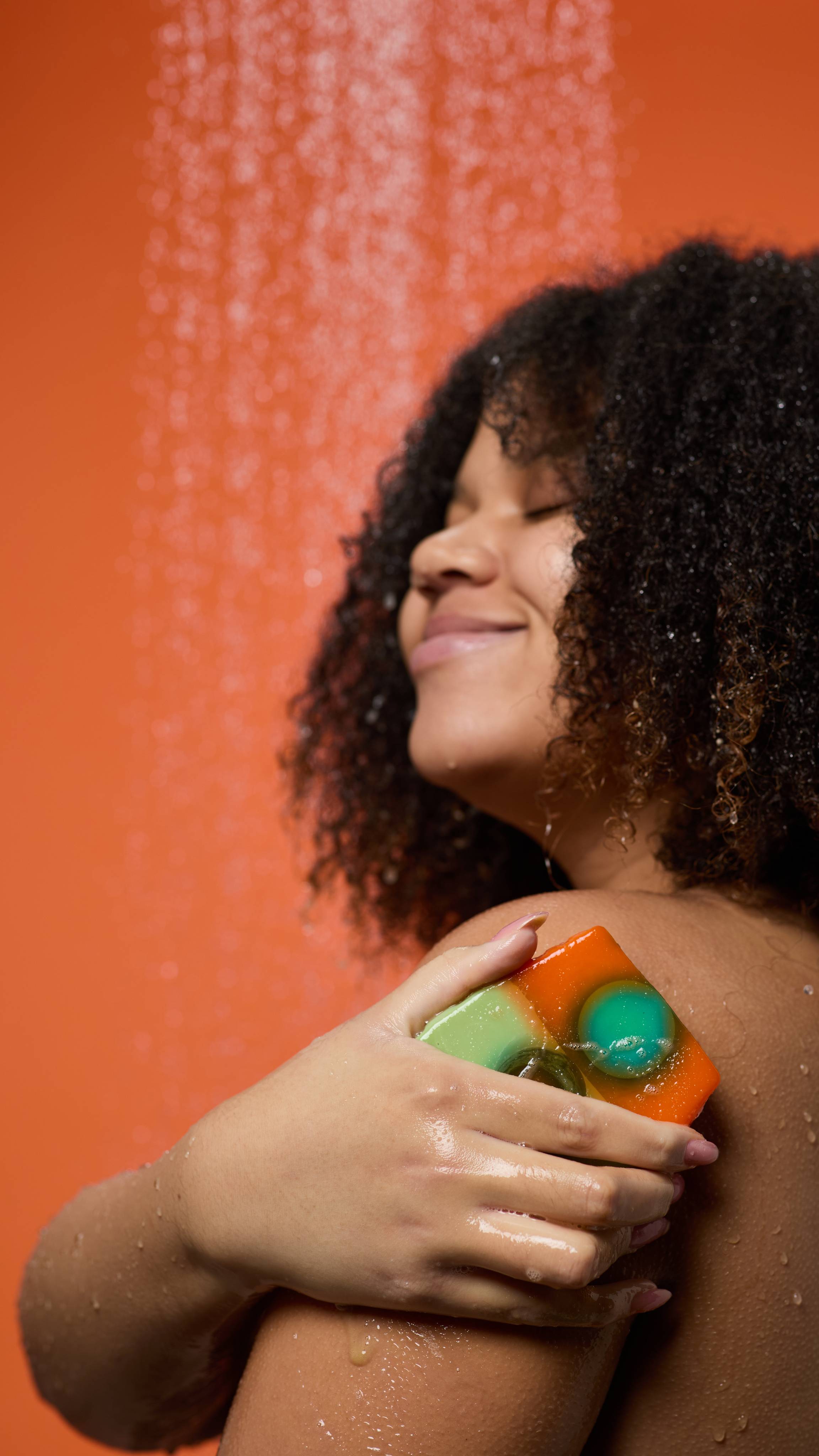 A close-up of the model's shoulder and facial profile, smiling, as they lather their upper arm with the Alban Elfed soap on a deep orange background. 