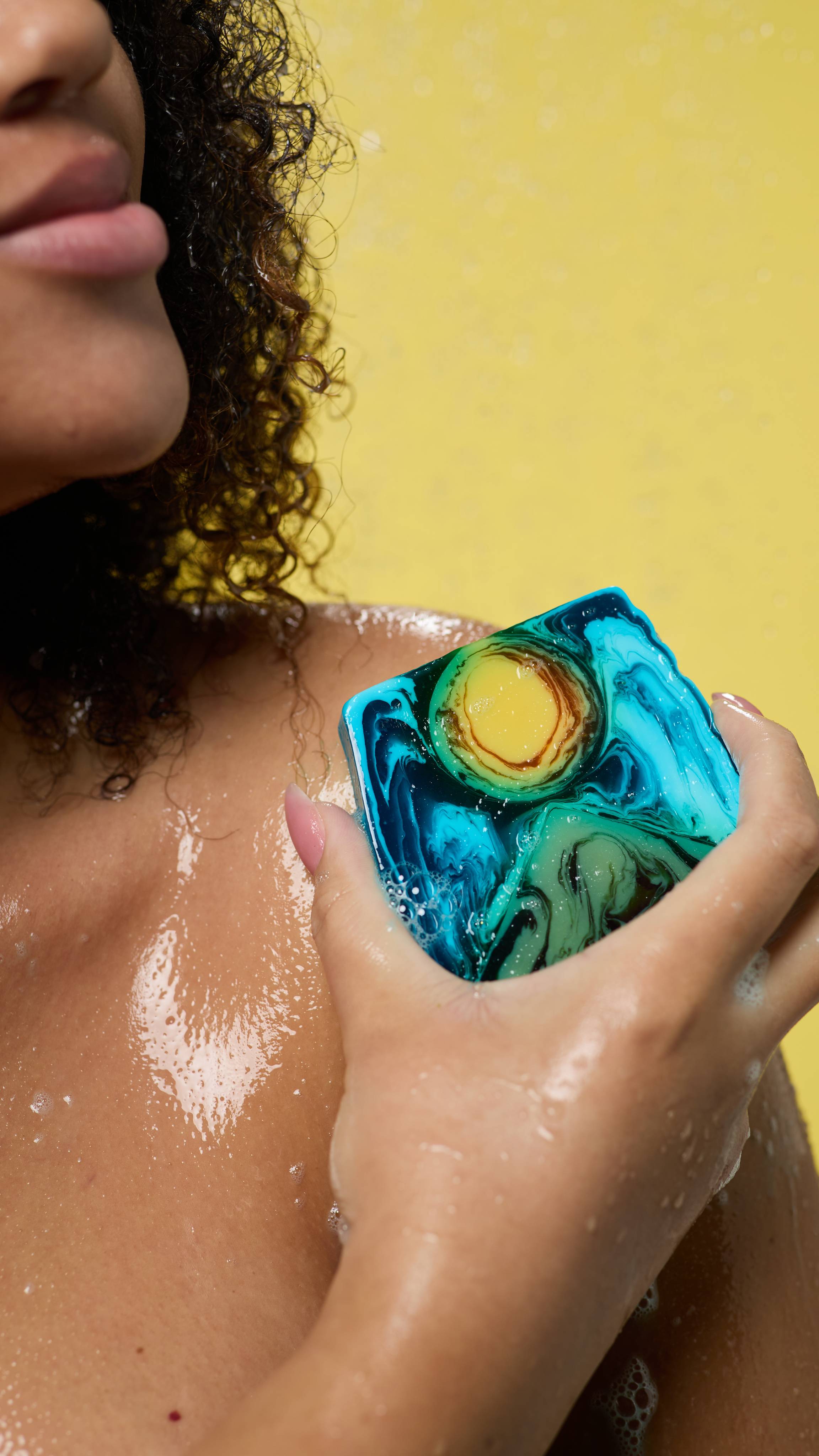 A close-up image of the model's hand holing the Alban Hefin soap in their hand as the smooth it over their shoulder under running shower water on a yellow background. 