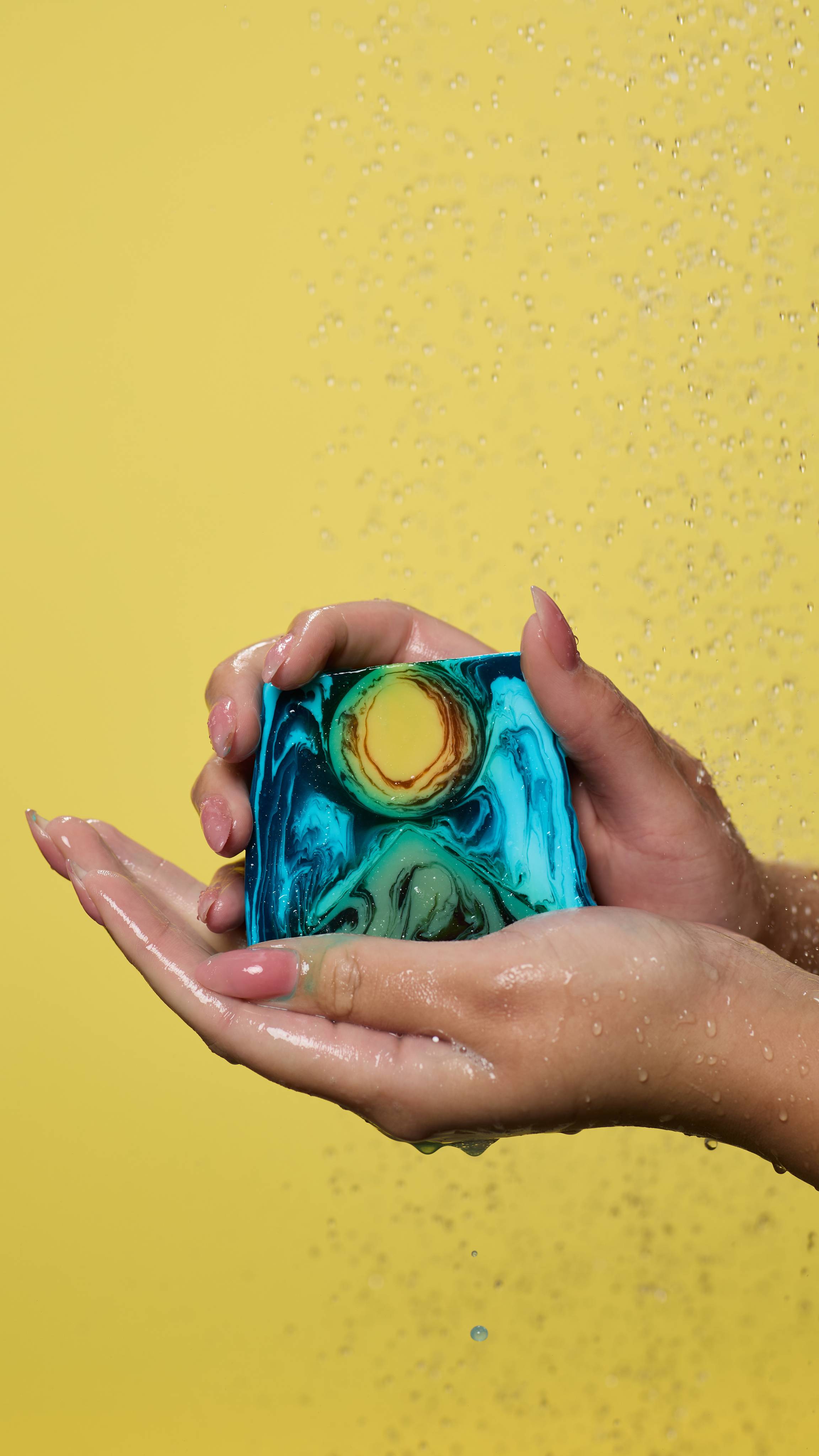 A close-up of the model's hands, lathered up with soapy suds while holding the Alban Hefan soap. Water is falling from above and there is a pastel yellow background.