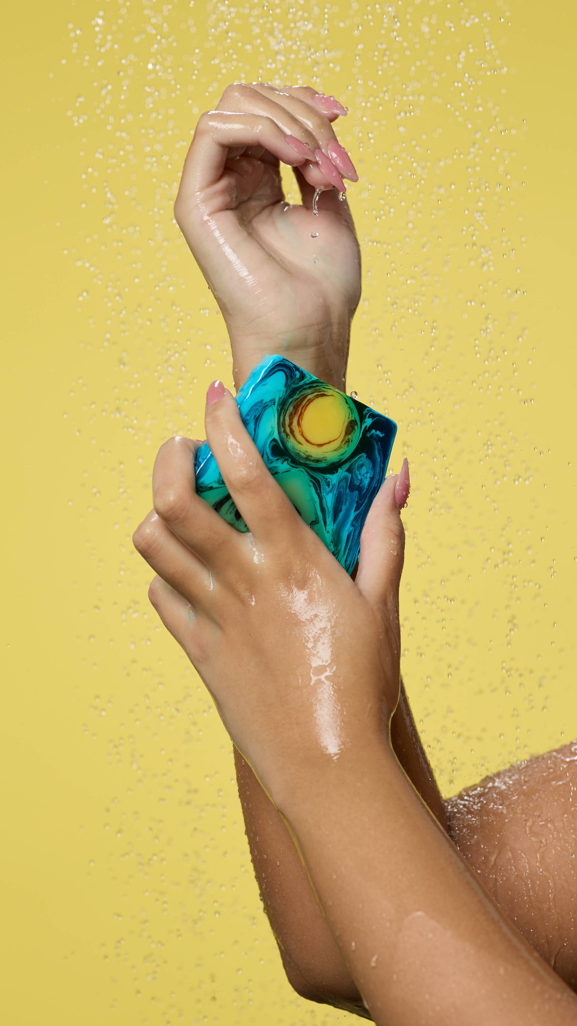 A shot of the model's arms on a yellow background as water is falling. They use one hand to smooth the soap over their forearm.