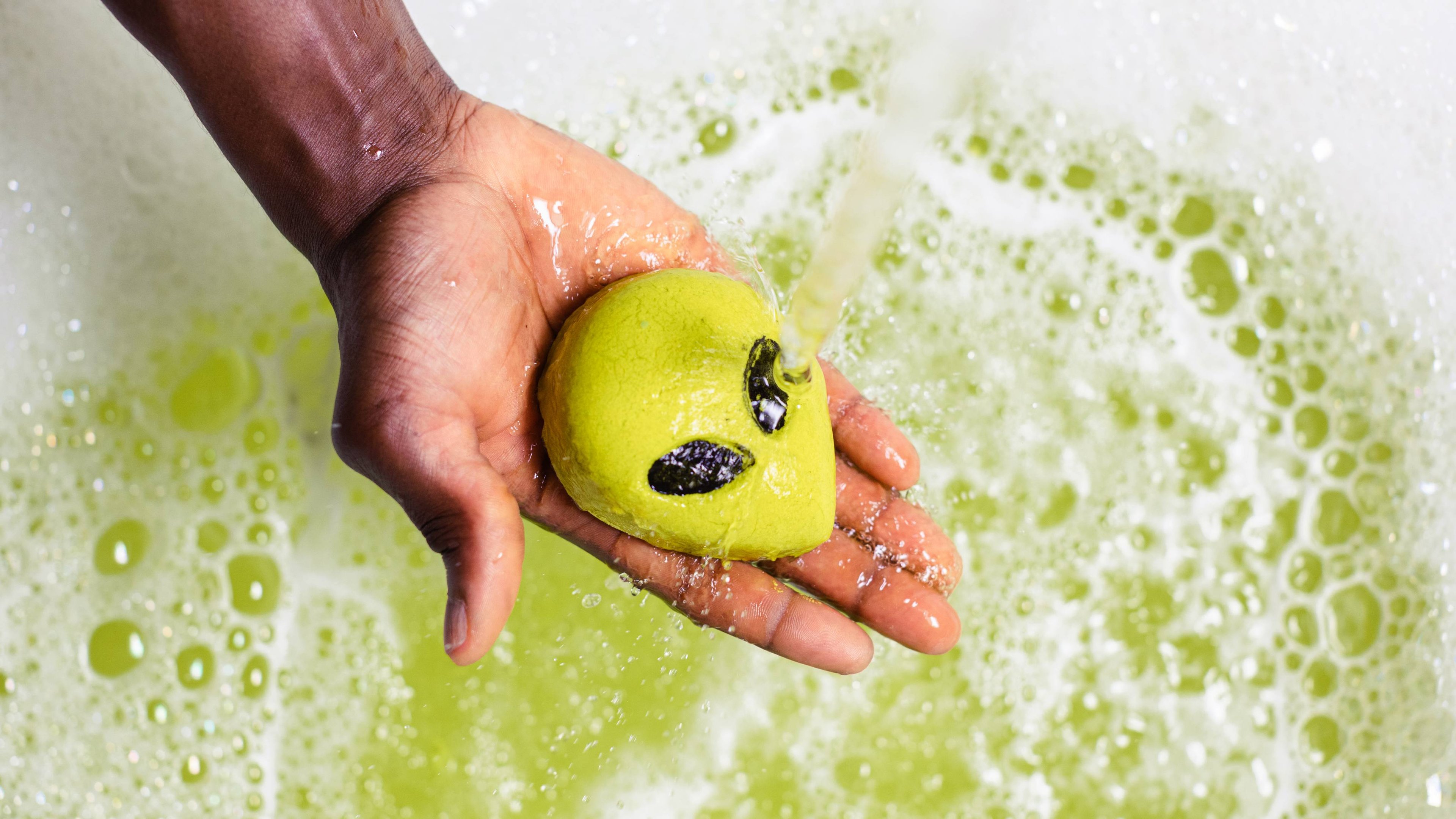 Model is holding the Alien bubble bar under running bath water above a foamy, bubble, ethereal-green sea of water.