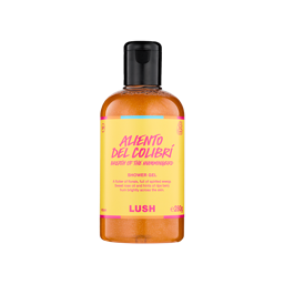A clear bottle filled with shimmering, golden-amber Lush Aliento del Colibrí shower gel, topped with a black cap and featuring a bright yellow label with pink text