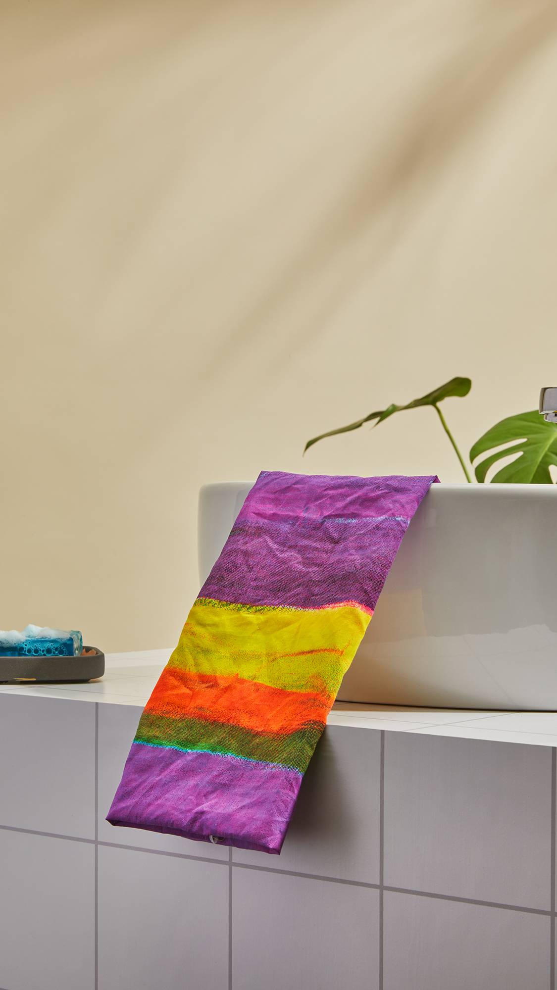 The image shows the colourful All The Colours Tenugui knot wrap folded neatly over the side of a sink as a freshly used soap slice sits beside it. 