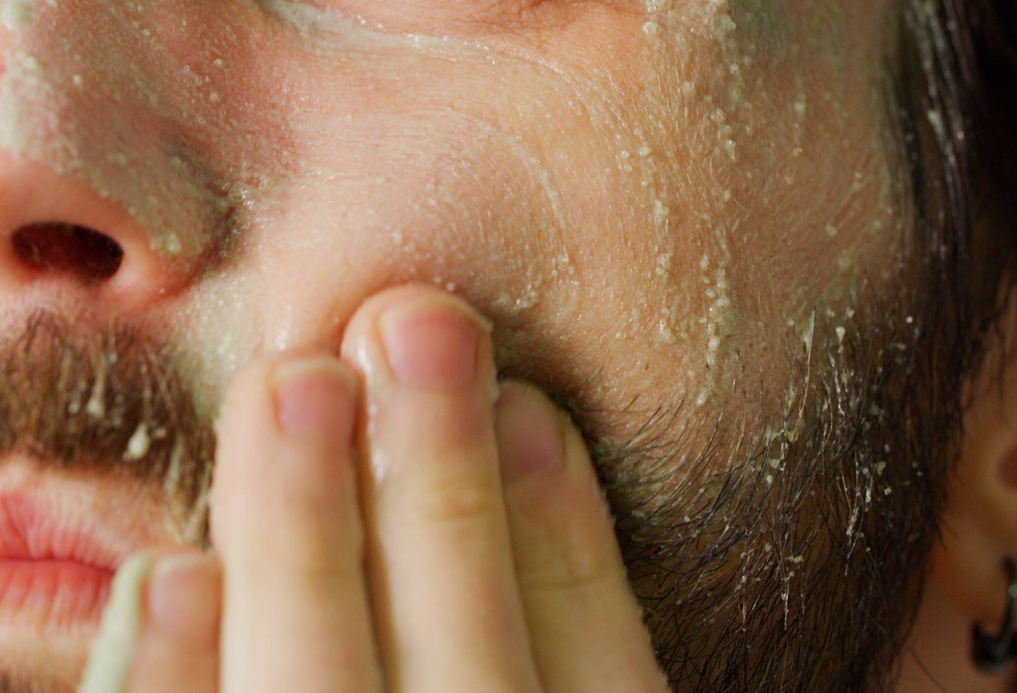 A bearded face is covered in a thin, light green paste with sugar crystals being rubbed into the lower cheek area.