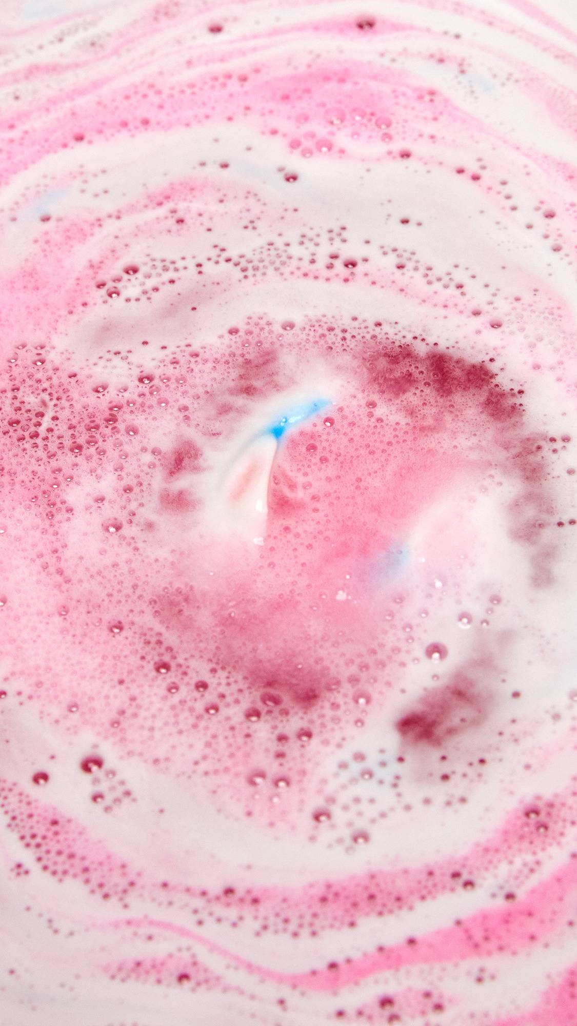 The American Cream bath bomb is slowly dissolving in the bath water giving off a pretty pink and white rippled foam across the surface with a small speck of blue in the centre.