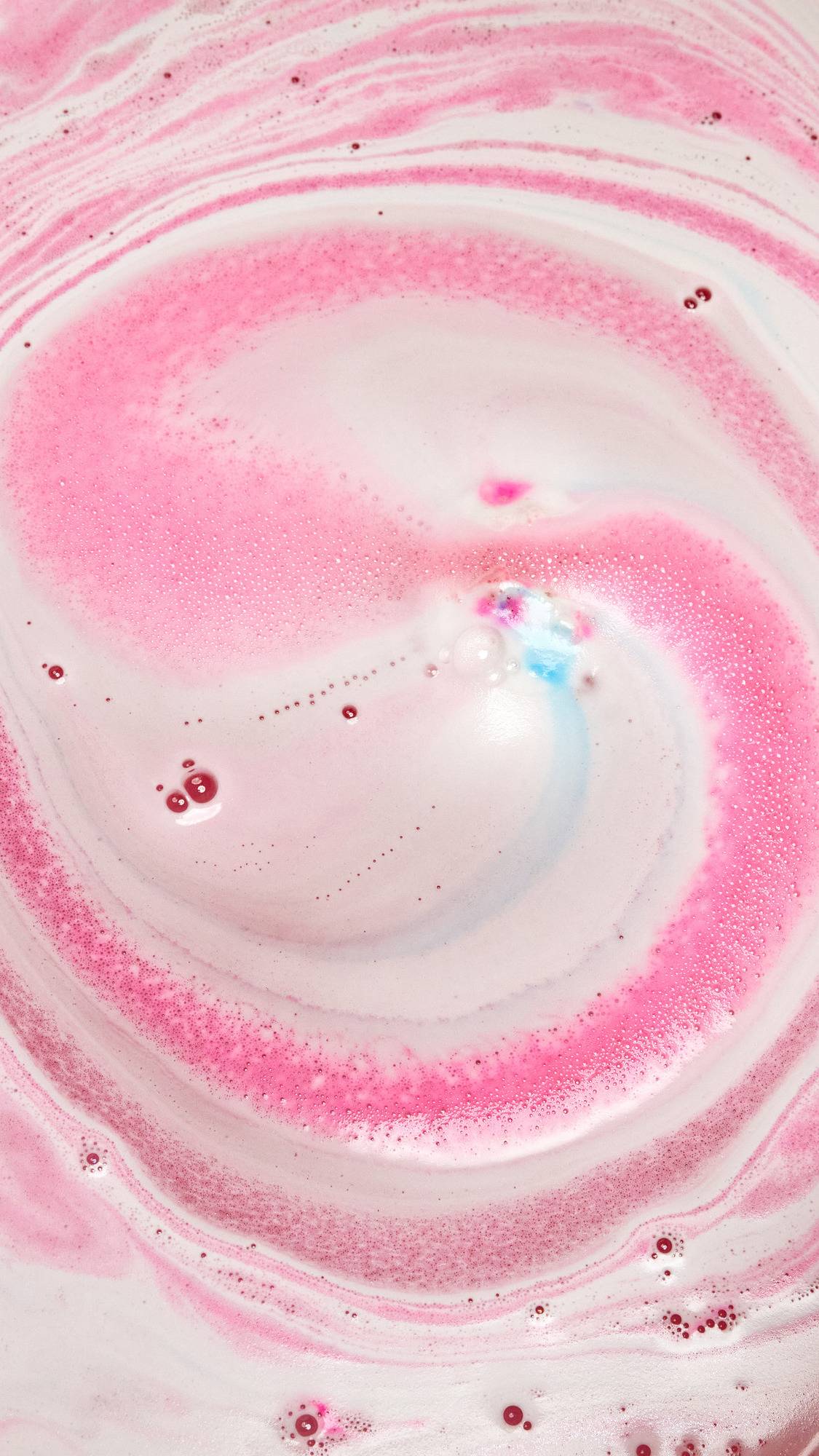 The American Cream bath bomb is slowly dissolving in the bath water giving off a pretty pink and white rippled foam across the surface with a small speck of blue in the centre.
