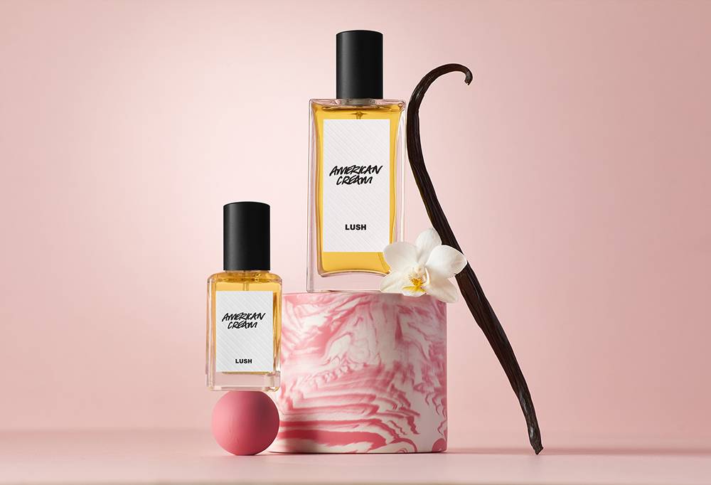 Two perfume bottles balance on a pink ball an cylindrical podium.  Next to it is a pink and white flower and a vanilla pod.