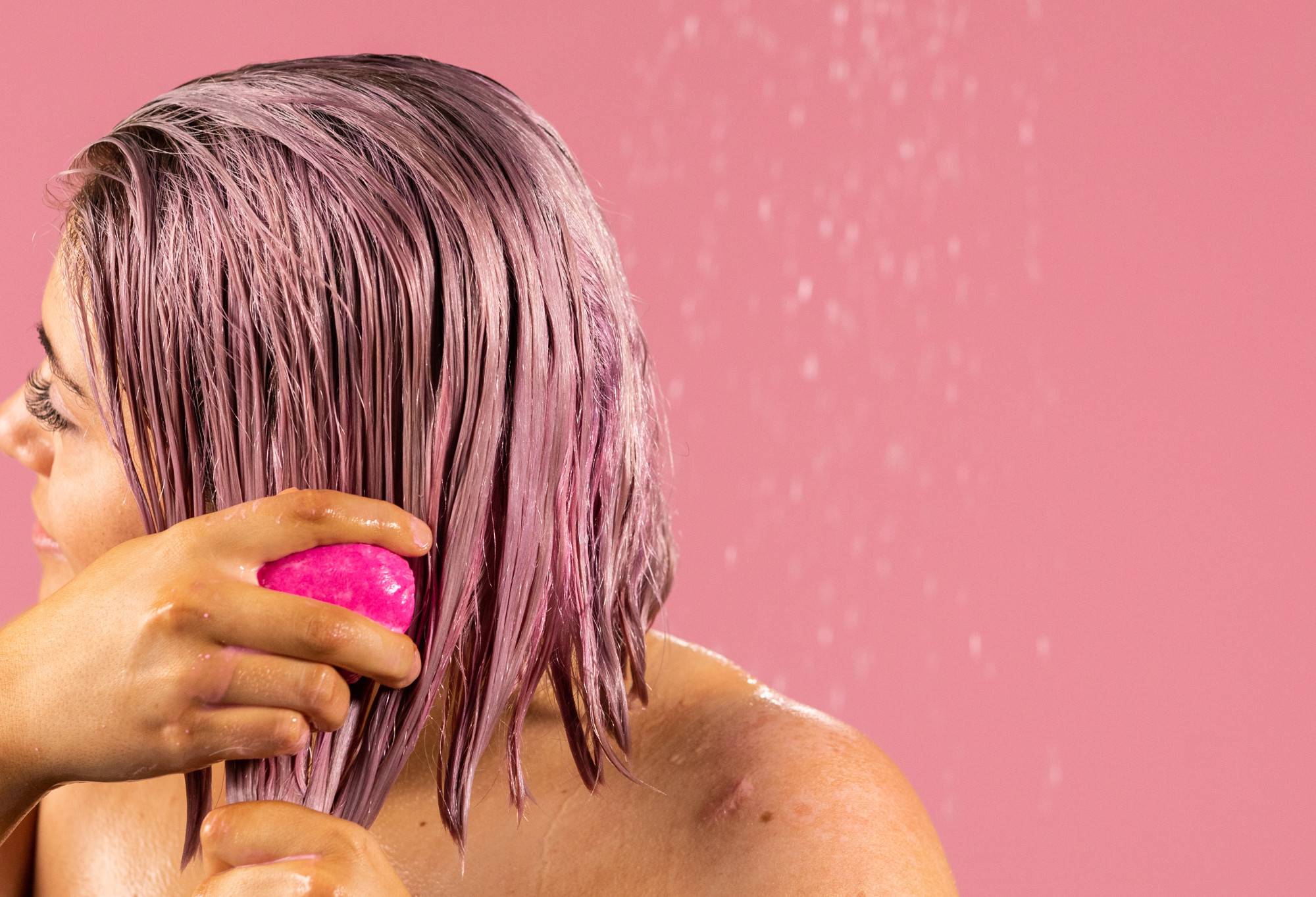 A person with bobbed pink hair works American Cream, a pink, oval shaped solid conditioner bar, through their hair.